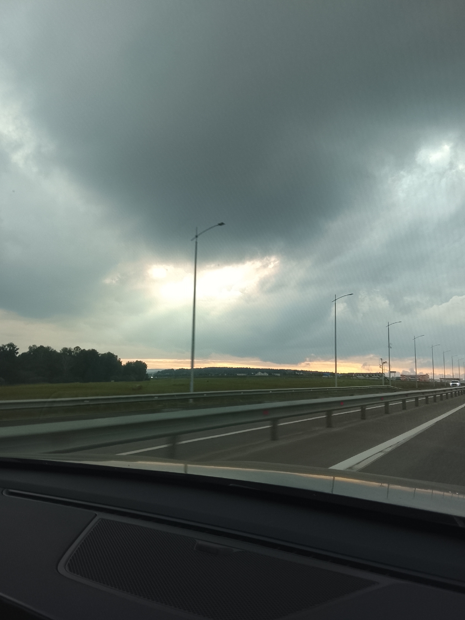 The image shows a view from inside a vehicle, looking out at a highway and a dramatic sky. The sky is filled with dark, heavy clouds, with a bright patch of sunlight breaking through in the middle distance. There are several tall streetlights lining the highway. On the left side of the highway, there is a line of trees, and beyond that, a flat landscape with some buildings visible. A white car is visible on the right side of the highway. The foreground shows the dashboard of the vehicle. The image appears to have been taken in the city of Элита, Russia.