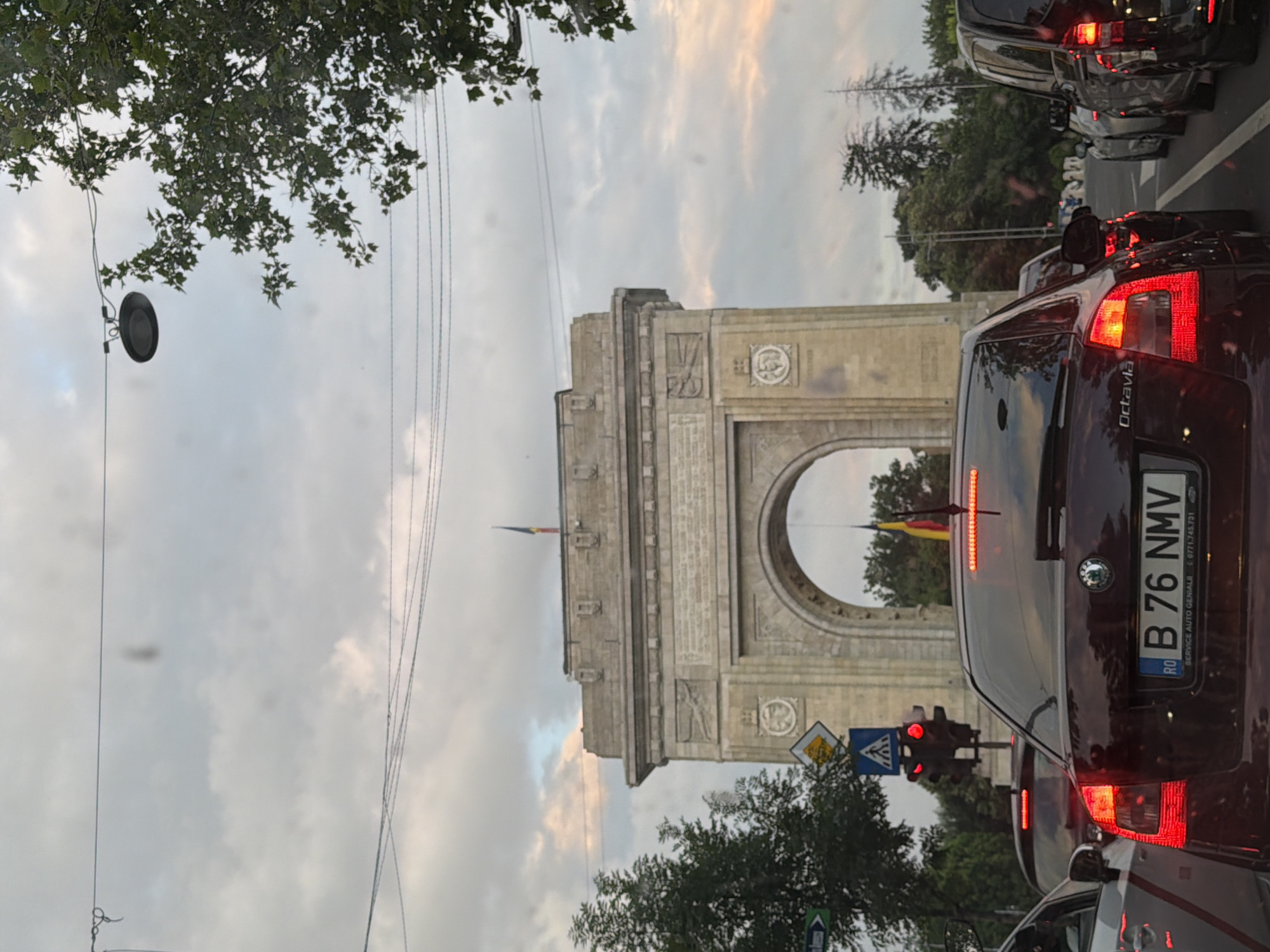 The image depicts a street scene in Bucharest, Romania, looking toward the Arcul de Triumf (Triumphal Arch). The foreground is dominated by the rear of a dark red/maroon Skoda Octavia car, facing away from the viewer, with its brake lights illuminated red. The Romanian license plate "RO B 76 NMV" is clearly visible. To the immediate left, a portion of a white car is visible, and to the right, the rear of another dark vehicle is present.
Traffic appears stopped, indicated by the red illumination of a vertical traffic light pole positioned on the left side of the road. Below the traffic light, a blue square sign with a white pedestrian symbol and a yellow diamond-shaped sign are mounted on a pole.
In the midground, beyond the cars, stands the Arcul de Triumf, a large, light-colored stone monument featuring a prominent archway. A Romanian flag, with vertical blue, yellow, and red stripes, flies from the top of the arch. The arch itself displays architectural details, including reliefs and decorative elements on its façade.
The sky above is predominantly overcast with grey clouds, though hints of lighter, possibly sunset-tinged clouds with pink and orange hues are visible on the horizon, particularly behind the arch. Utility wires and cables stretch across the upper portion of the sky. A dark circular street light fixture hangs from wires on the upper right, partially obscured by green tree foliage. Additional green trees line both sides of the road and frame the monument in the background. No people are discernible in the scene.