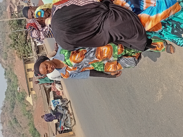 The image portrays a daytime street scene in Ogaminan, Nigeria. A paved road occupies the foreground, extending into the midground where it is bordered by buildings and individuals. In the center-left foreground, a young adult female stands, looking directly at the camera. She wears a black headscarf, a light blue collared shirt with green, orange, and grey abstract patterns, matching patterned trousers, and long black undersleeves. Dark sandals are visible on her feet. To her right, the back of another individual, likely female, is partially visible, wearing a black head covering and a vibrant blue, orange, and green tie-dye patterned dress or wrapper.
Behind these two figures, a small group of people is present. One person wears a black cap, and another has a multi-colored patterned headwrap (red, black, white). On the left side of the midground, a silver motorcycle, equipped with a blue and white patterned box on its rear seat, is parked. A man in dark blue attire stands near the motorcycle, facing away from the viewer. Further back, a man in a green sleeveless top and trousers is visible near buildings. Several large white sacks are resting on the ground in this vicinity. The background features residential-style buildings with light-colored walls and brown corrugated roofs, situated at the base of arid, sparsely vegetated hills under a clear sky.