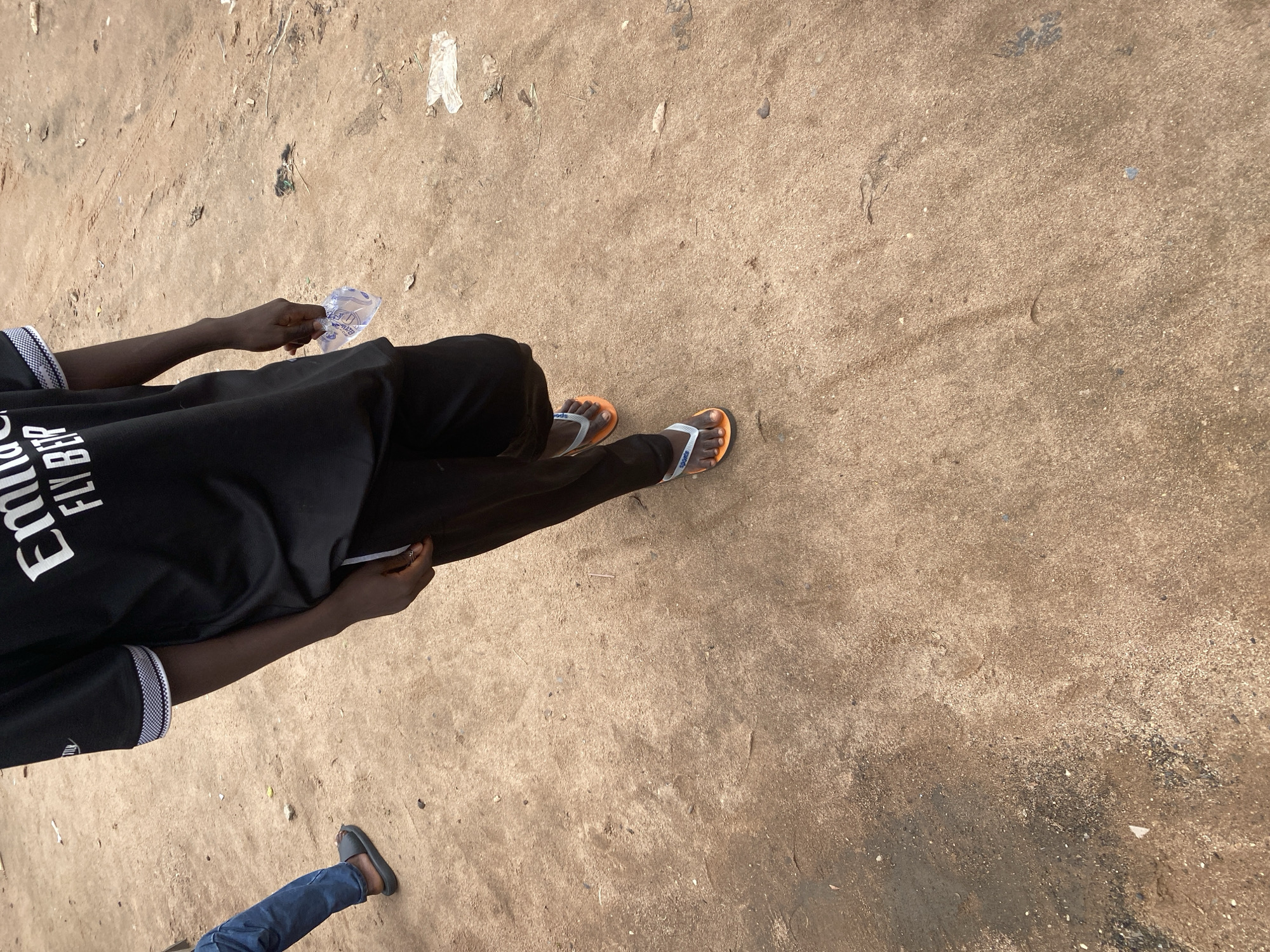 This is a medium shot, taken from a high angle, of two individuals in Jalingo, Nigeria. The primary subject is a person wearing a black t-shirt with white lettering and black pants. They are holding a small, clear plastic bag in their right hand. Their feet are clad in orange and white flip-flops. The person is standing on a dusty, light brown ground that appears to be a road or pathway.
To the lower left of the frame, another person's legs are visible. They are wearing blue jeans and dark gray slip-on shoes. This second person appears to be walking away from the camera.
The lighting suggests it is daytime, likely outdoors. The ground surface is dry and shows signs of wear, with small debris scattered across it. The overall impression is of an ordinary day in a somewhat arid environment. The t-shirt worn by the primary subject has visible white text that appears to say "EMIRATES FLY BETTER."