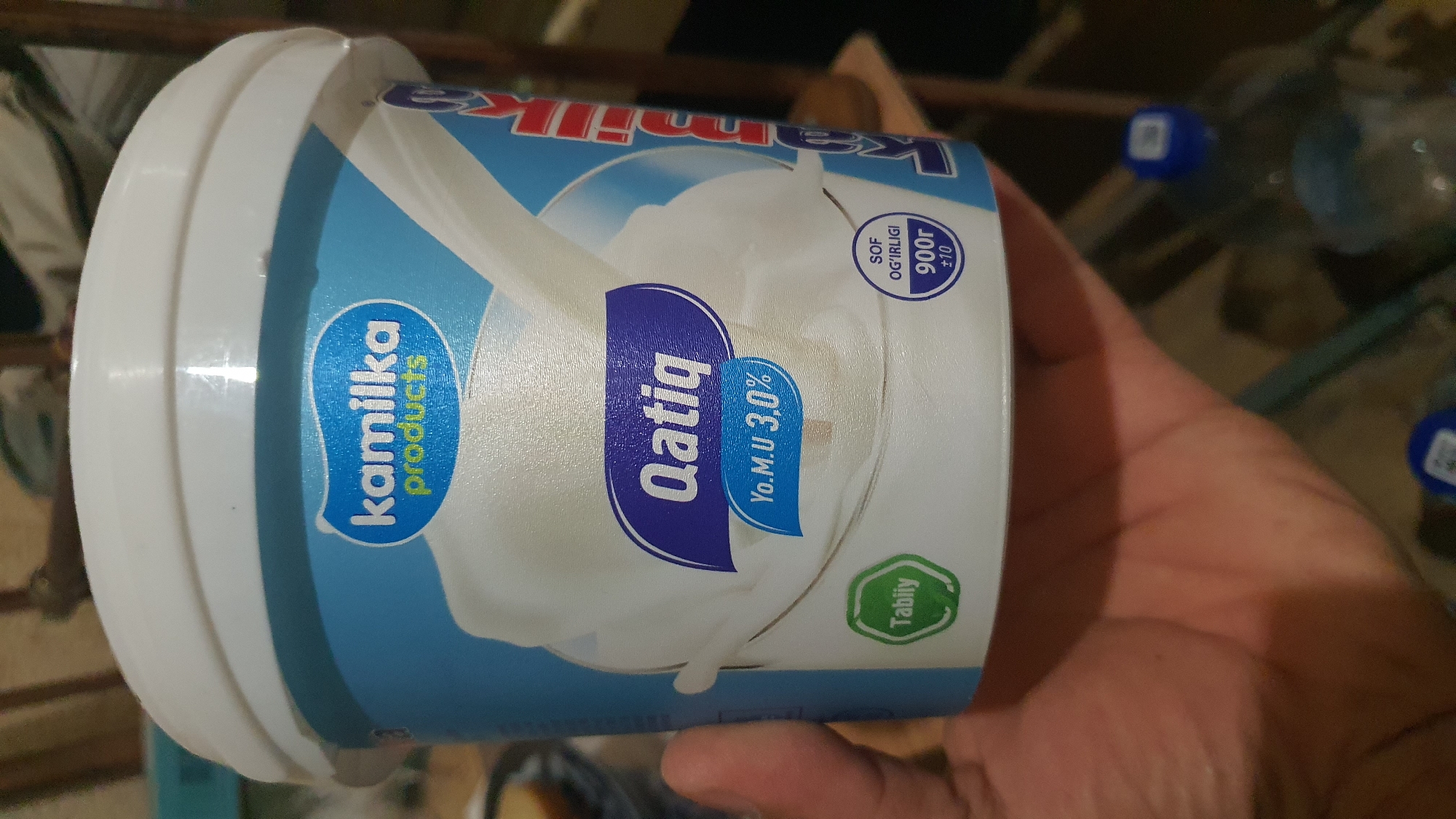 A person is holding a white plastic tub of "Qatiq" (yogurt) with a blue and white label in Karankul, Uzbekistan. The label features the brand "Kamilka Products" in blue and white lettering. The tub indicates "Yo.M.U 3,0%" (fat content) and "Sof Og'irligi 900r ±10" (net weight). A green oval sticker with "Tabiiy" (natural) is also visible on the tub. The background is blurred, showing indoor elements possibly in a home or store setting.