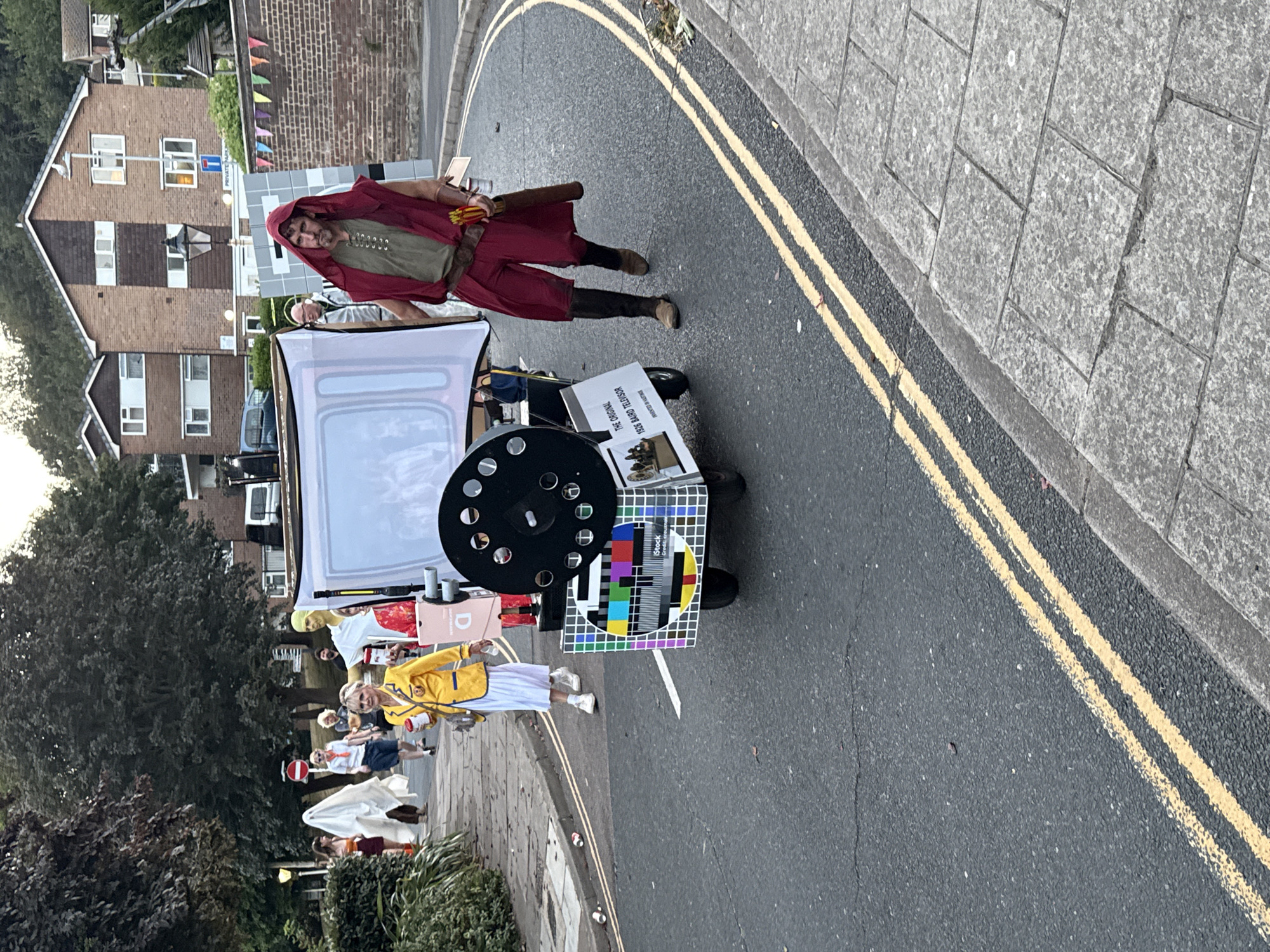 In St Leonards, United Kingdom, on a residential street with a paved sidewalk and asphalt road, a parade is underway. The focus is on a cart decorated to resemble an old television set, complete with a test card pattern on its screen. A man in a red hooded robe and brown boots walks alongside the cart, holding what appears to be a quiver of arrows. Behind him, a woman in a yellow blazer and white skirt carries a drink. Other individuals, some in costumes like a ghost and a person with a yellow head, are visible in the background, participating in the event. Buildings line the street, and trees and foliage are present. The weather appears to be overcast, contributing to a muted lighting condition.