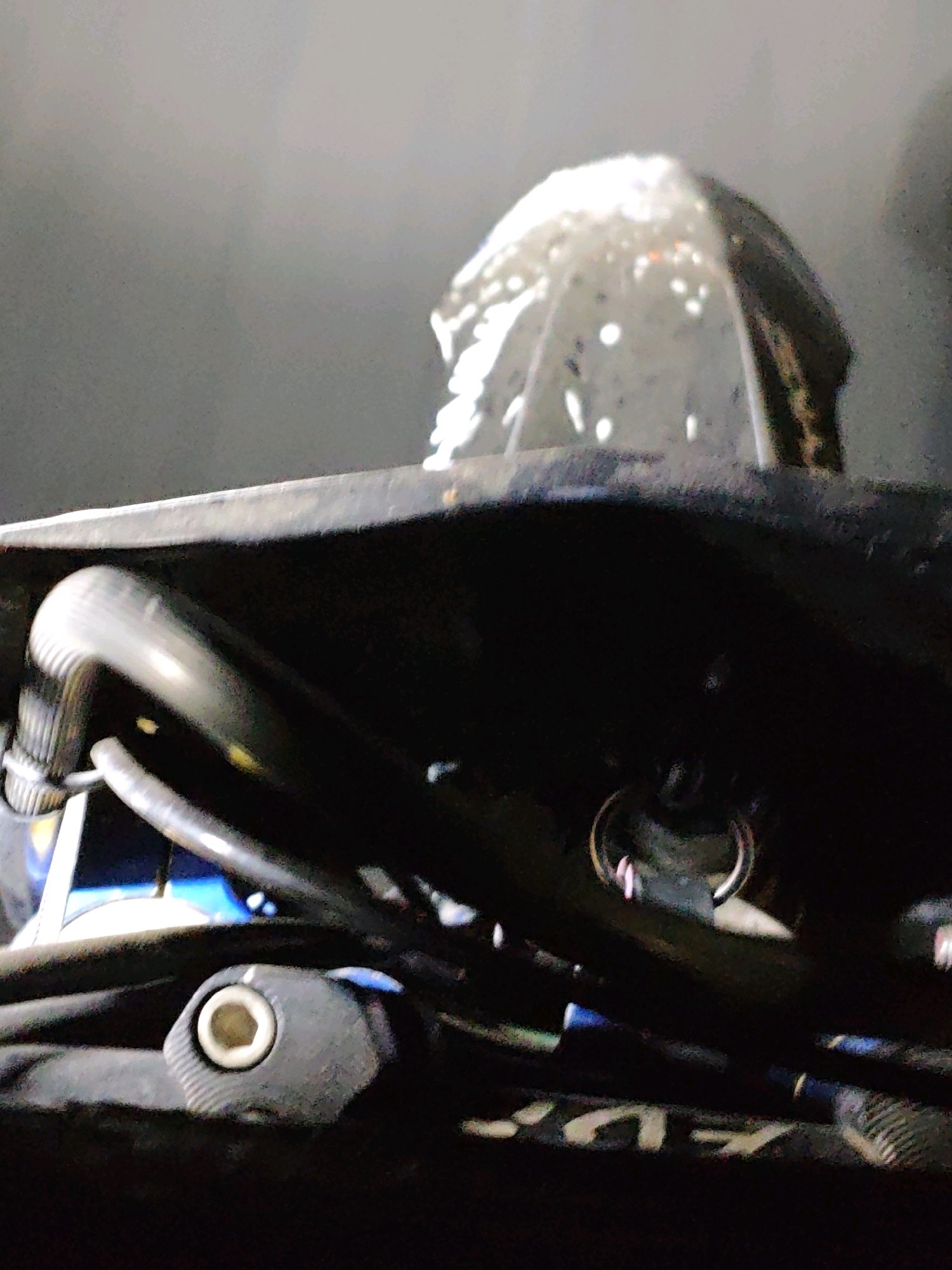 The image shows a close-up, low-angle shot of a piece of scuba diving equipment, possibly a regulator or a part of a dive computer. The equipment is predominantly black with some metallic accents, including a silver ring visible towards the center. There are black hoses and tubes, and a part with a hexagonal indentation. The top of the equipment is obscured by a dark, curved surface. A transparent or translucent dome-like object with white speckles is visible on top of this surface. The background is a muted, dark grey, suggesting an indoor or dimly lit environment. The lighting highlights the texture and metallic sheen of the equipment. The image does not depict any discernible location within the city of Muara Dua, Indonesia; it is a close-up of technical equipment.