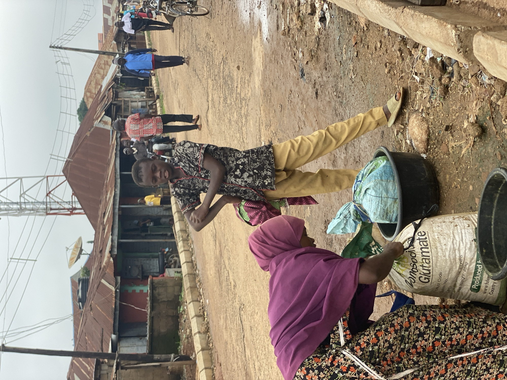 The image shows a street scene in Kafanchan B, Nigeria. A young person wearing a dark patterned shirt and yellow pants is standing and smiling at the camera. Another person, wearing a purple head covering and a dark floral-patterned dress, is bending over a large white bag with "Monosodi Glutamate" printed on it. Next to the bag are two black buckets. In the background, other people are walking along the dirt street. There are buildings made of corrugated metal and a communications tower visible in the background. The ground is dirt and littered with debris.