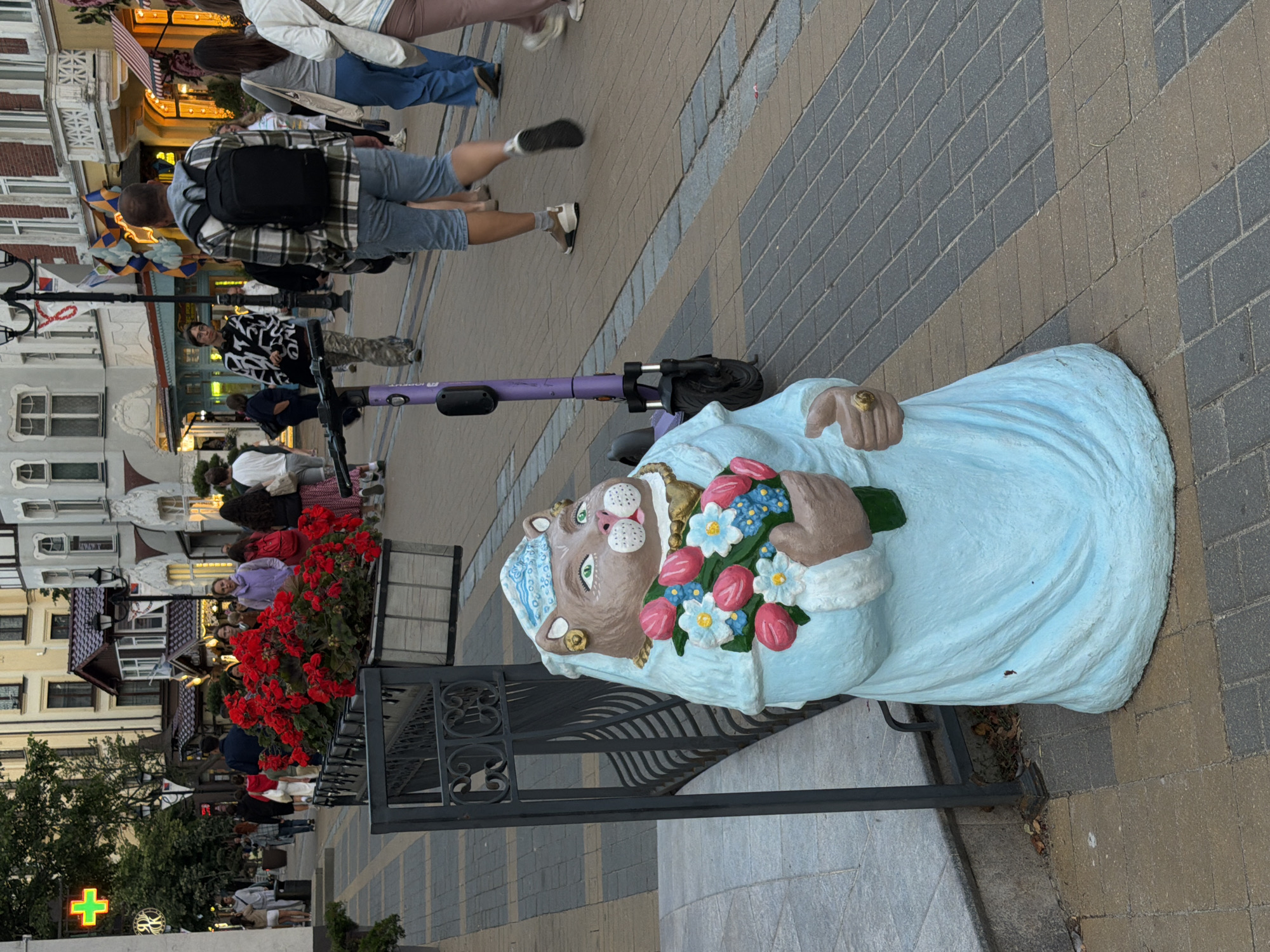 In Zelenogradsk, Russia, a statue of a cat wearing a blue dress and headscarf holds a bouquet of tulips and daisies. The cat statue, situated on a brick sidewalk, is adorned with a gold ring on its right paw. To the left of the statue, a metal bench and a planter filled with red flowers are visible, while a purple electric scooter stands nearby. Pedestrians walk along the street, passing by shops with awnings. A green cross sign indicates a pharmacy in the background, and trees line the sidewalk.