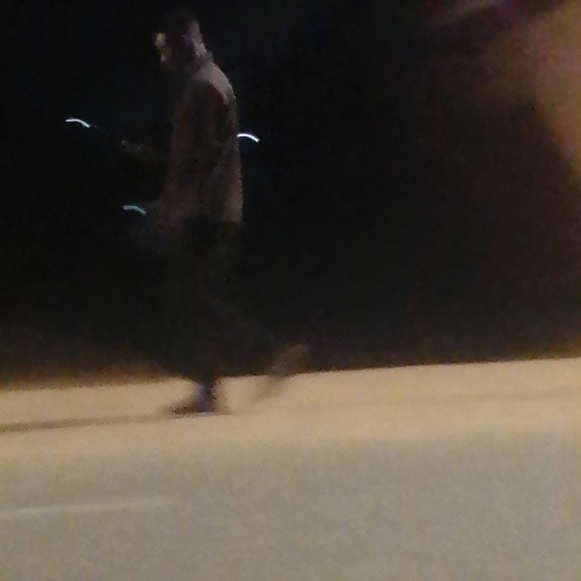 A person is walking on a paved surface at night. The person is wearing a dark jacket and pants. The background is dark and blurry, with some faint lights visible. The paved surface appears to be a sidewalk or street. The image is grainy and low resolution, making it difficult to discern fine details. The overall impression is of a solitary figure moving through a dark urban environment.