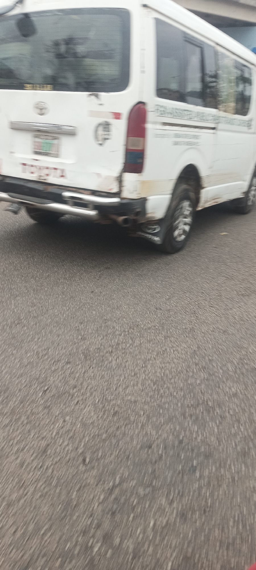 The image shows the rear of a white Toyota Hiace minivan driving on a paved road. The van is dirty, with mud splattered on its lower panels and rear bumper. A chrome bumper guard is installed, and dual exhaust pipes are visible. The license plate is partially obscured but appears to have green characters. The van has a rear window with a wiper. Text and logos are visible on the van's body, including a Toyota emblem and what appears to be an inscription on the side. The scene is set in the city of Kabong, Nigeria, characterized by the worn asphalt road and the type of vehicle commonly used for public transportation or commercial purposes in the region. The image is slightly blurred due to motion.
