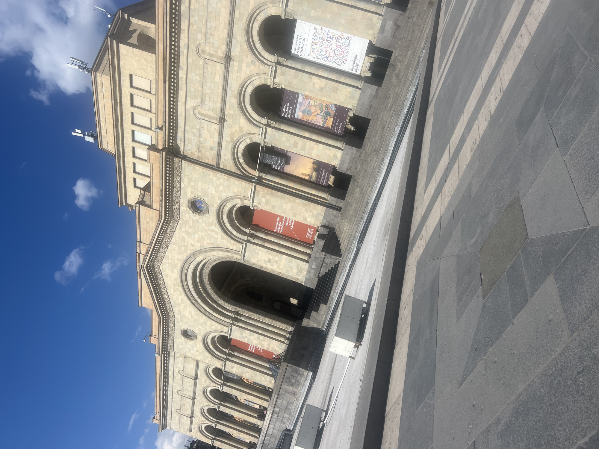 The image shows the National Gallery of Armenia in Yerevan. The architecture is visible with its facade and arched entrances. Banners promoting exhibits are hung between the arches. The sky is clear blue with some clouds, and the foreground is a gray stone-tiled area.