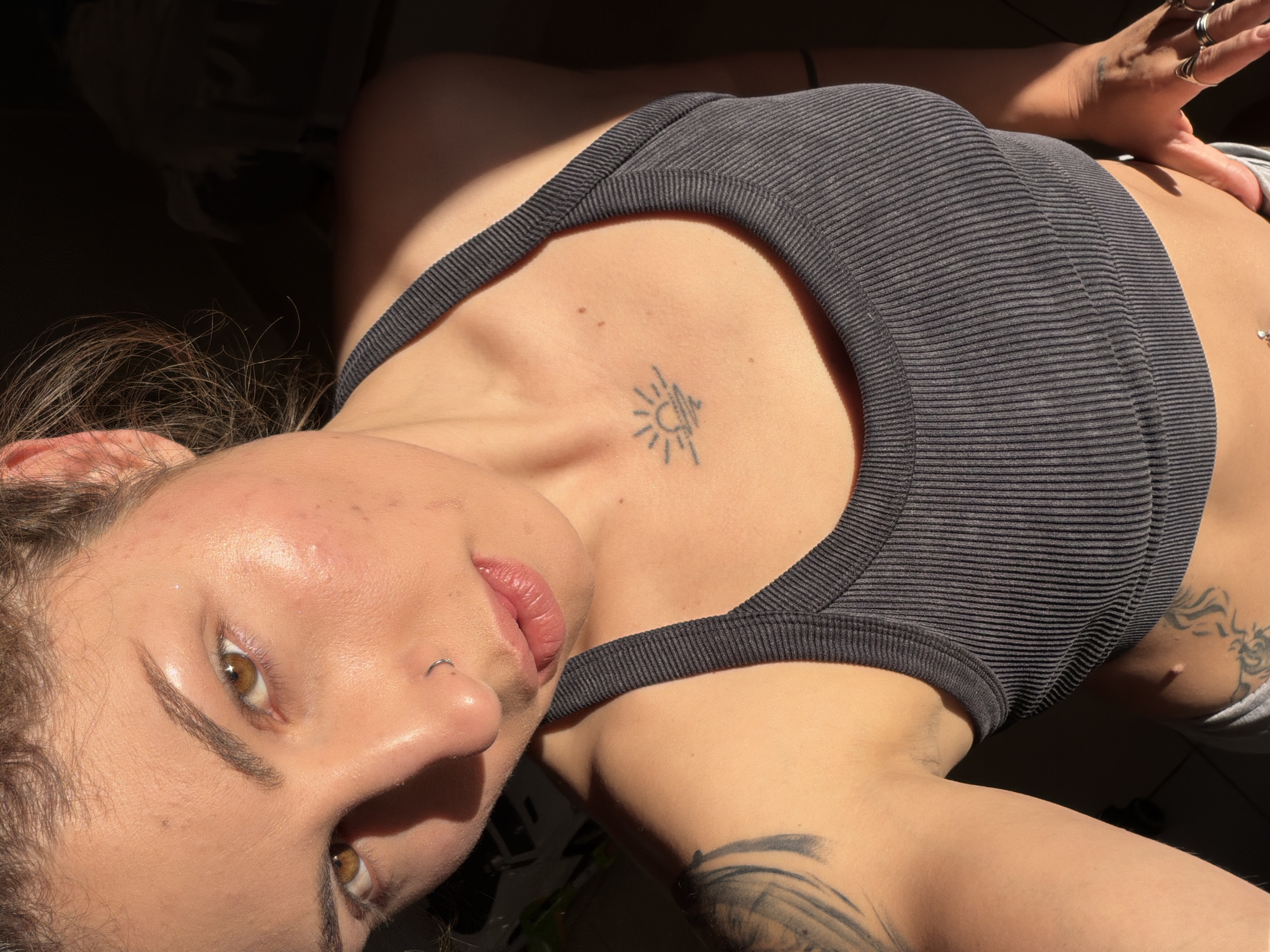 A young woman is shown in a close-up, angled, eye-level shot. She has fair skin and light brown hair. Her eyes are hazel-colored, and she has a nose piercing. She is wearing a gray ribbed tank top. A tattoo of a sun is visible on her chest. Another tattoo is partially visible on her arm. The lighting is bright and casts shadows across her face and body. No location information in the city of Kiryat Bialik, Israel is visible in the image.