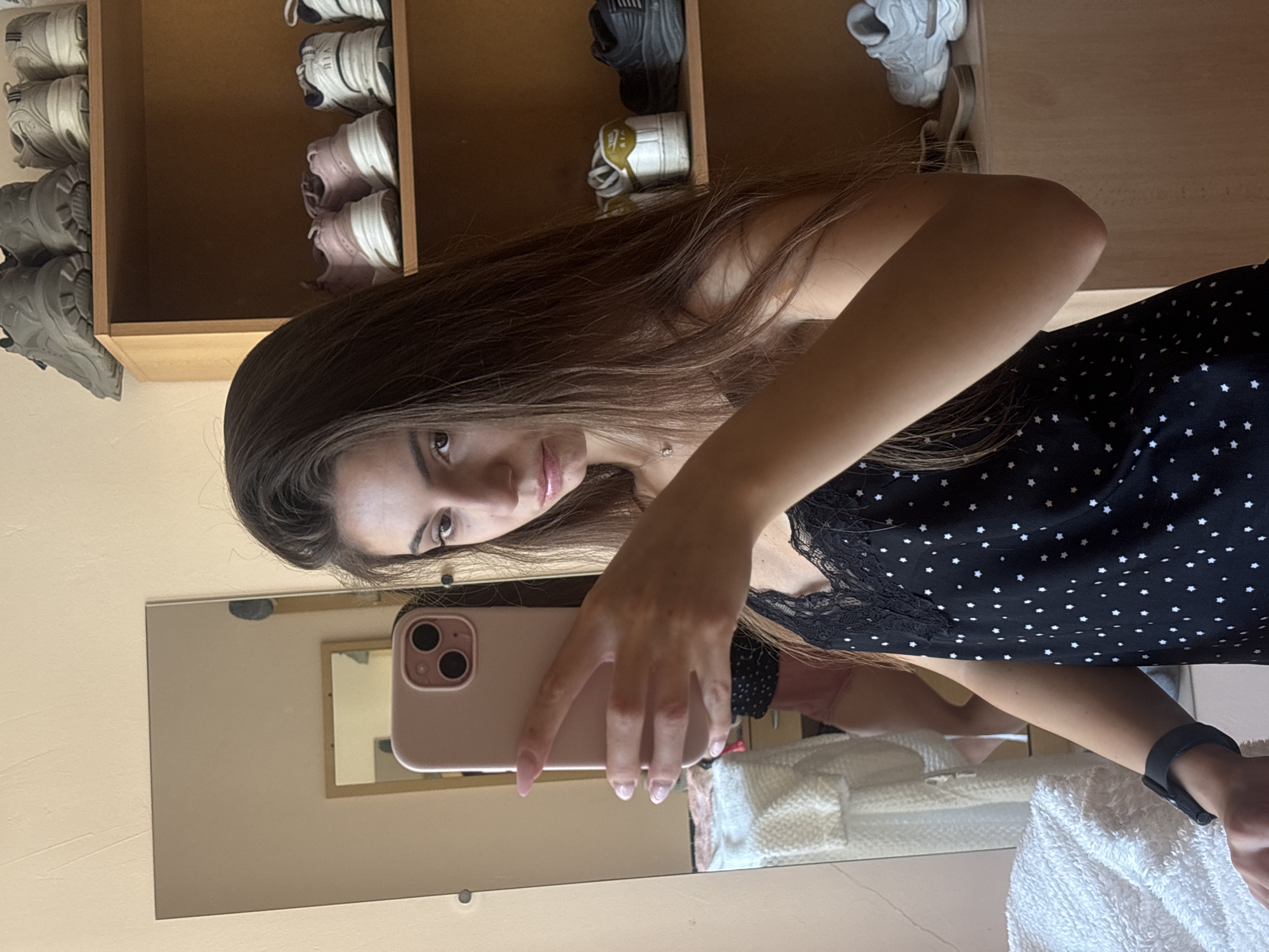 In Varna, Bulgaria, a young woman with long, dark brown hair takes a mirror selfie in what appears to be a bedroom. She is wearing a black sleeveless top with a white star pattern and a black lace trim around the neckline. She is holding a pink smartphone in her left hand, pointing it towards the mirror. Her right hand is partially visible, resting near her chest. In the background, a wooden shelf unit is visible, filled with various pairs of sneakers. To the left of the woman, a white fluffy blanket or towel is draped over a surface, and a large mirror reflects a small framed mirror and a blurred image of what seems to be clothing in the background. The wall behind the woman is a pale, textured color.
