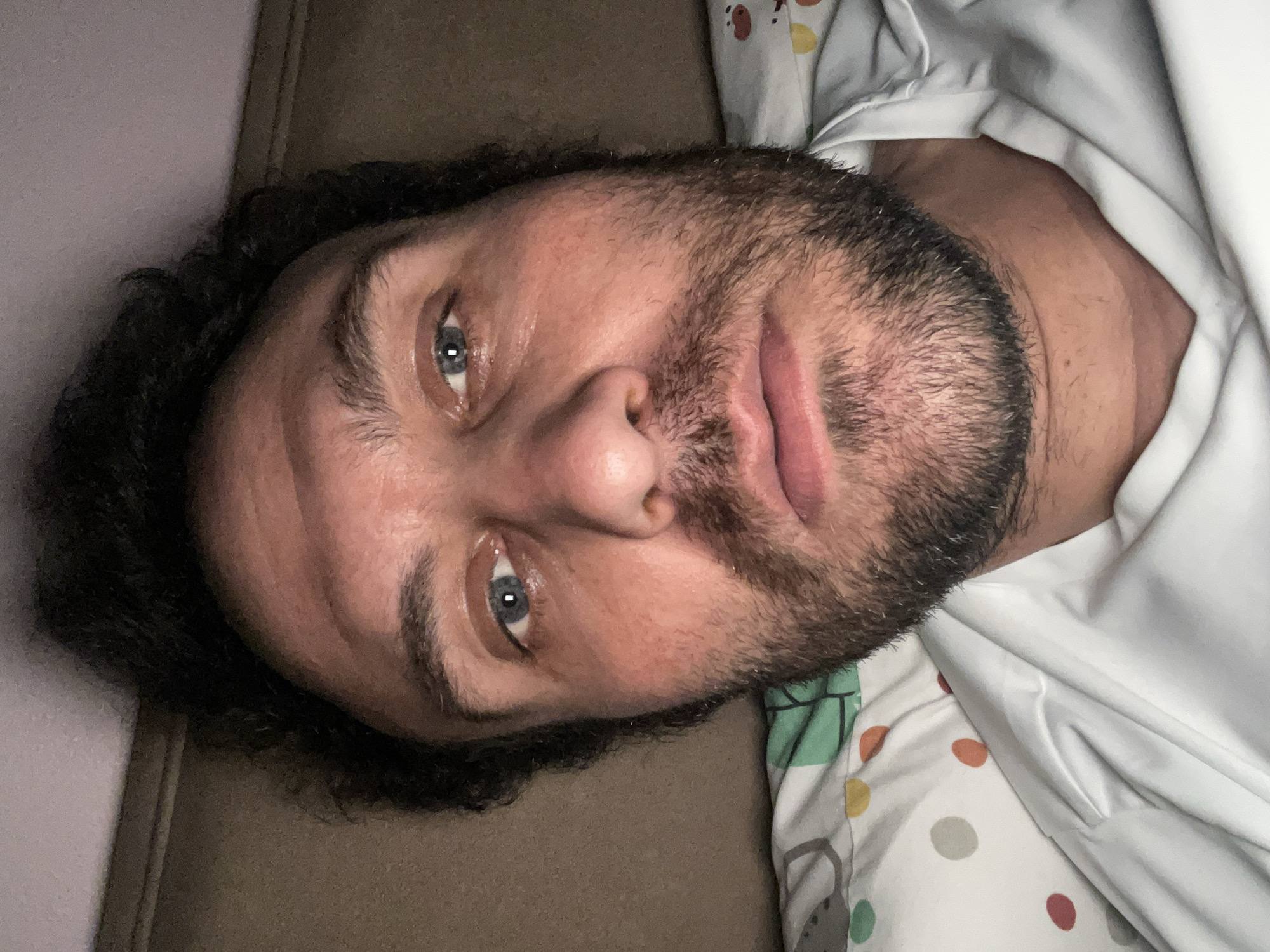 A medium close-up shot depicts a man with fair skin, dark curly hair, and a beard, resting his head on a brown surface, potentially a couch or pillow. He has blue eyes and a neutral expression. He appears to be wearing a white shirt with a pattern of colorful polka dots and cartoon images. The background includes a light purple wall. There is no evidence to suggest this image was taken in Armagh, United Kingdom.
