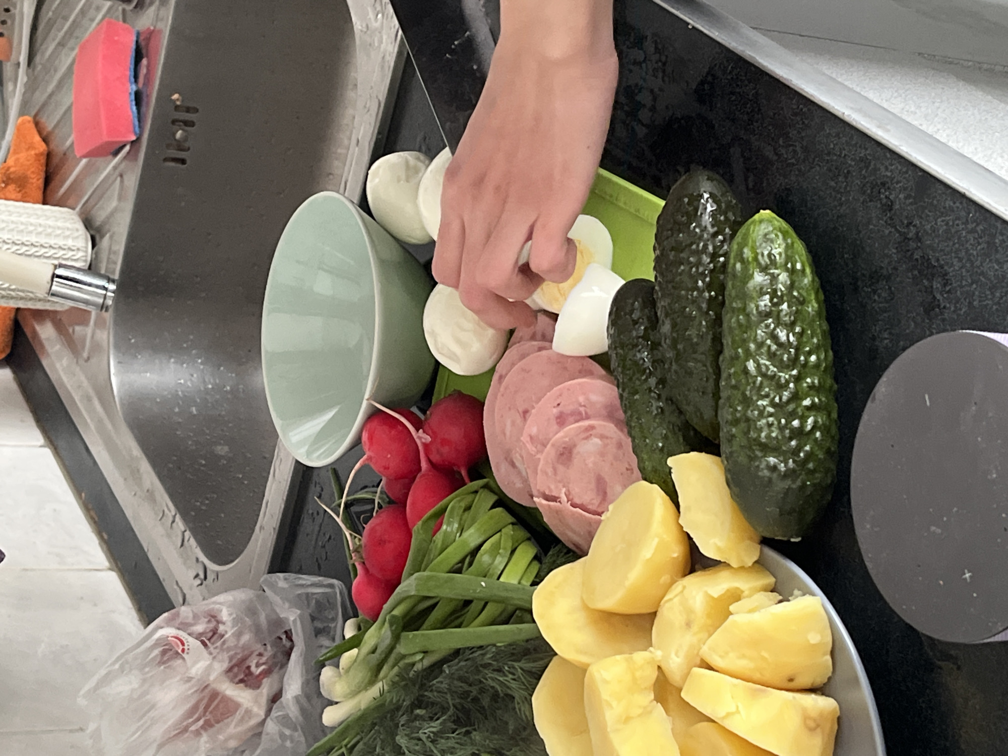 The image shows a kitchen scene with ingredients for a salad or similar dish. A hand is present, near sliced boiled eggs. Other visible ingredients include cucumbers, sliced sausage, radishes, green onions, dill, and boiled potatoes. The ingredients are arranged on a kitchen counter near a sink. The location is presumed to be a kitchen within an unknown city and region.