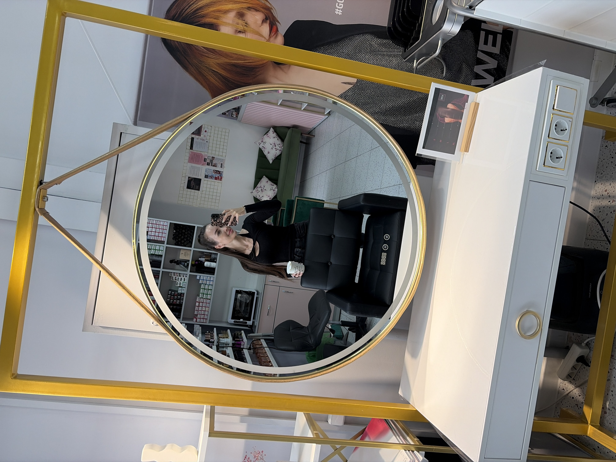 The image shows a woman taking a selfie in a round mirror, which is framed by a large yellow metal structure. The mirror reflects a salon interior with shelves filled with hair products, a black salon chair, a green sofa, and a microwave. On the desk in front of the mirror, there are electrical outlets and a small framed picture. The woman is wearing a black top and jeans, and she is holding a cup. In the reflection of the mirror, a large poster of a woman with orange hair is visible on the wall. The scene appears to be a modern hair salon. The question mentions Saint Petersburg, Russia, but there are no specific visual cues in the image that confirm this location.