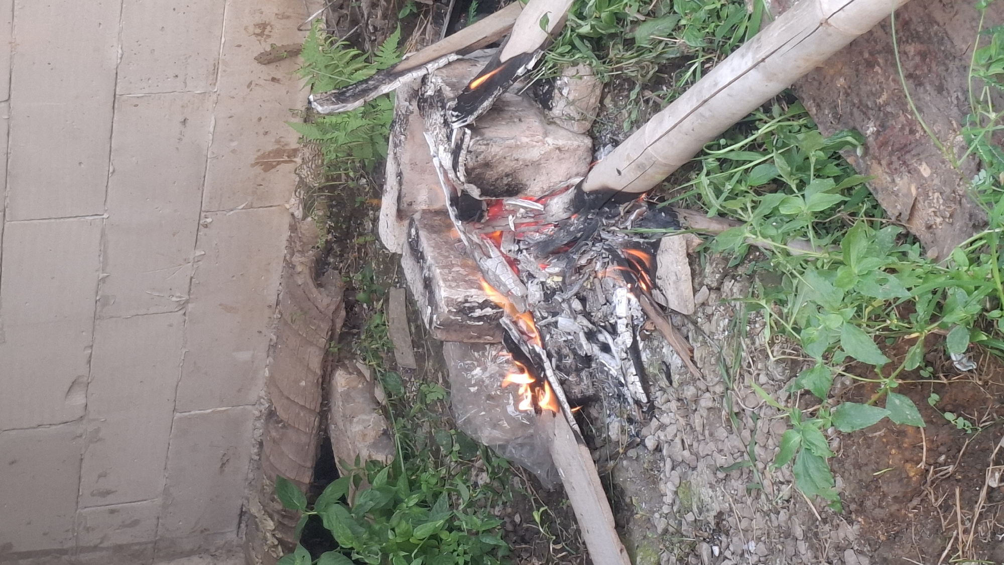 A fire burns in an outdoor setting in Rahong, Indonesia. The fire is contained within a makeshift structure of bricks. Several pieces of wood are ablaze, with embers and ashes scattered around the fire pit. The ground is covered with gravel and dirt, with some green vegetation growing around the edges. In the background, a wall made of concrete blocks is visible. The scene appears to be in a natural or semi-natural environment, possibly a backyard or a rural area. There are no people visible in the image.