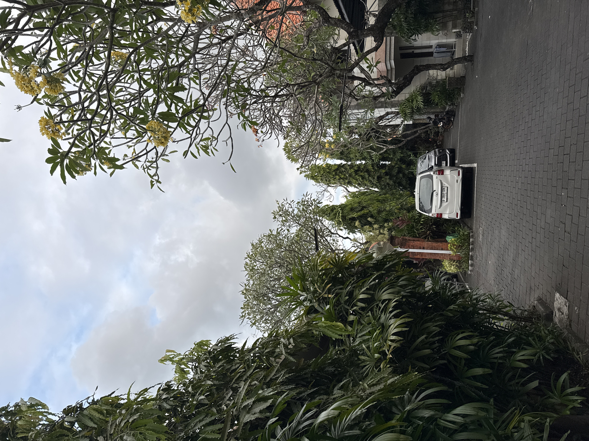 The image depicts a paved driveway lined with lush green foliage and trees, some bearing yellow flowers, leading towards a building with a terracotta-tiled roof. Two cars, a white minivan and a dark-colored SUV, are parked on the driveway. A motorcycle is also visible, parked near the building. The sky is overcast with scattered clouds. The scene appears to be within a residential or hospitality complex in Denpasar, Indonesia, given the typical architectural style and vegetation.