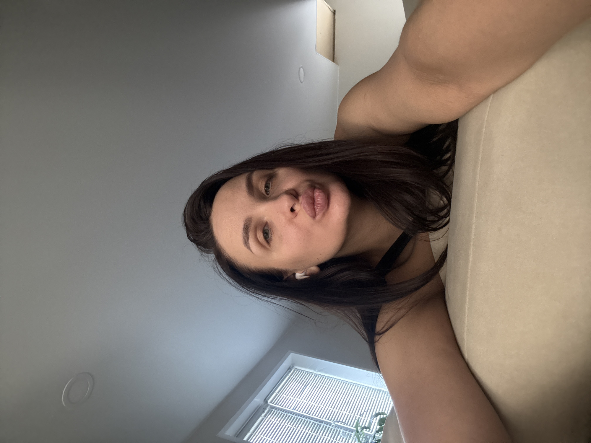 A woman with dark hair and light eyes is taking a selfie indoors. She has an earbud in her left ear. She's wearing a dark top. A window with blinds is visible in the background. Part of a light-colored couch is also visible. The location is not discernible.