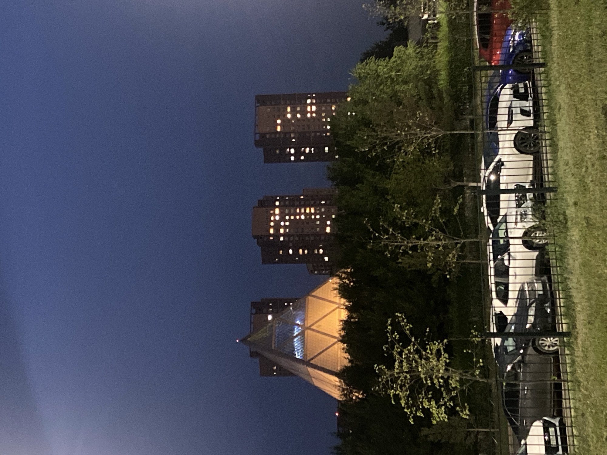 A nighttime exterior shot shows a modern architectural landscape in Astana, Kazakhstan. In the background, a unique pyramid-shaped building, possibly a cultural or administrative center, is illuminated with warm yellow light, its glass apex reflecting the dim sky. To the right of the pyramid are two tall, rectangular residential buildings, their numerous windows glowing with warm light, indicating occupancy. The buildings are surrounded by a dense canopy of dark green trees, which obscure the ground level in the distance. In the foreground, a dark metal fence runs horizontally across the image. Beyond the fence, several cars are parked in a lot. Visible cars include a dark sedan, several white SUVs and sedans, a dark blue sedan, and a red sedan. The ground in front of the fence is covered in short, green grass. The sky is a deep, dark blue, suggesting twilight or early night.