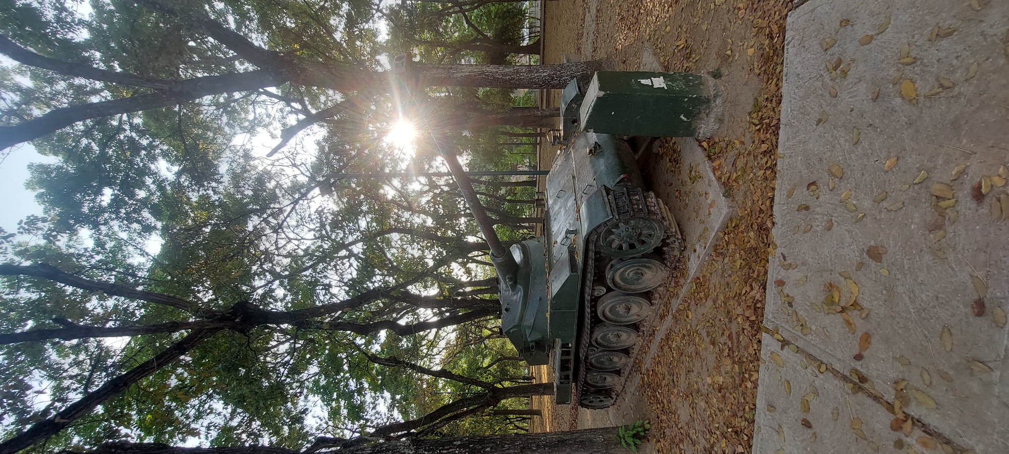 The image depicts an old, green tank on display in a park. The tank is positioned on a concrete base surrounded by fallen leaves. Trees with dense foliage overhang the tank and the surrounding area. The sun is shining brightly through the trees, creating a glare. The location is identified as Caracas, Venezuela. No people are visible in the image.
