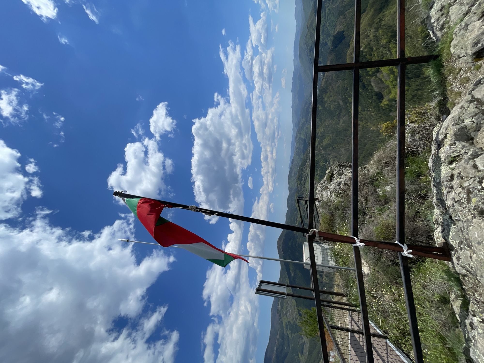 A vertical shot captures a panorama from what appears to be an elevated viewpoint in Gradat, Bulgaria, featuring a Bulgarian flag prominently displayed against a backdrop of a partly cloudy blue sky.
The flag, positioned to the left of the frame, is secured to a horizontal pole. The colors are green, white, and red, top to bottom.
To the right, a metal railing borders the edge of the vantage point, offering a view of rolling hills and valleys. The landscape stretches into the distance, with varying shades of green indicating vegetation. The sky transitions from a light blue at the horizon to a deeper blue overhead, punctuated by fluffy white clouds.