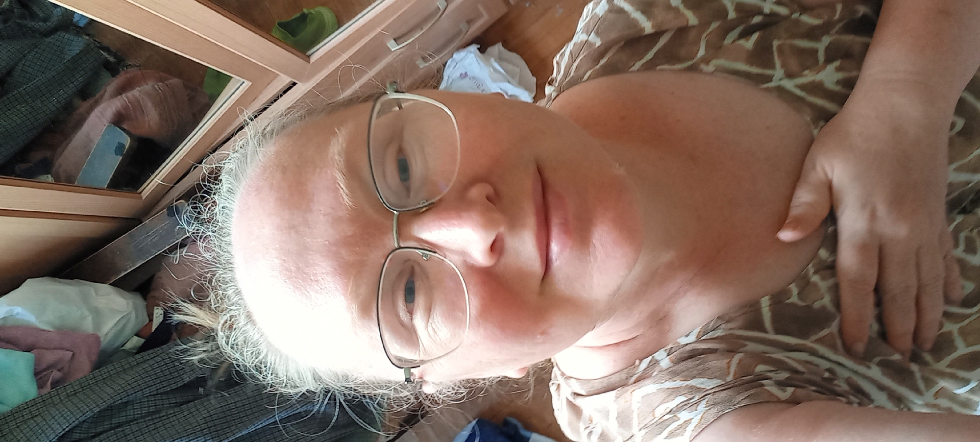 An overhead close-up selfie shows a Caucasian woman with fair skin and light-colored hair. She is wearing glasses with thin metal frames and a brown and white patterned sleeveless top. Her right hand is placed on her chest. The background is blurred and appears to be an indoor environment with wooden furniture and some items of clothing or fabric. The overall scene is a casual, personal photograph.