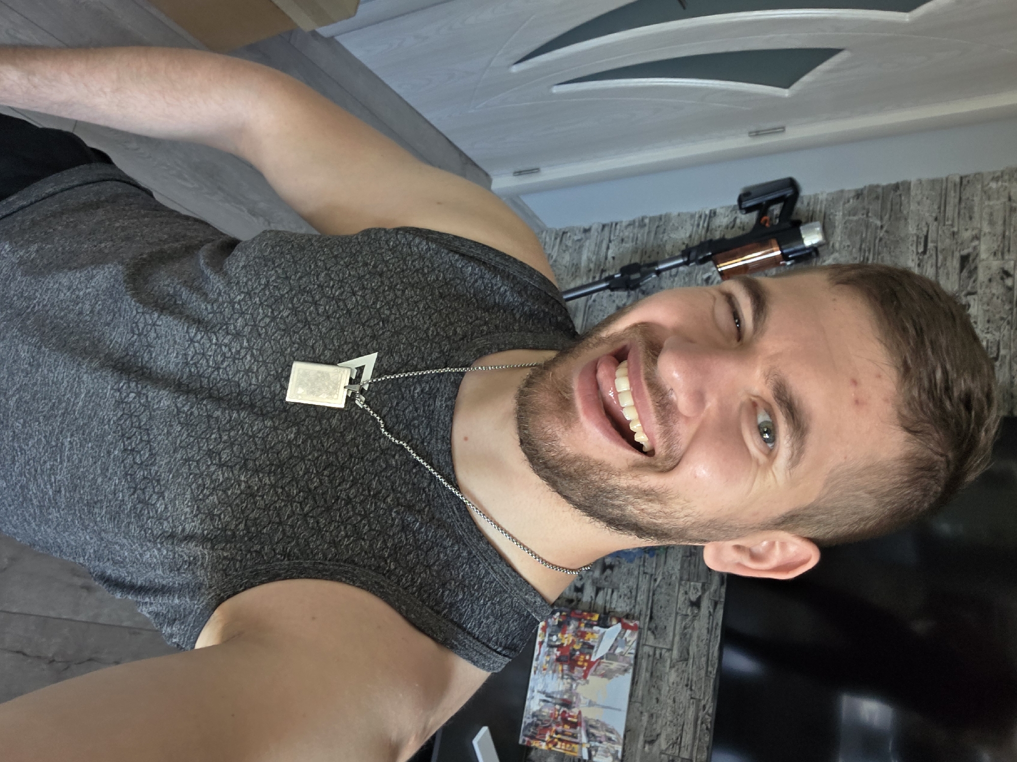 A man is positioned at an oblique angle in the frame, facing the camera and winking. He has short brown hair, a trimmed beard, and is wearing a grey tank top with a dog tag necklace. The background includes a white patterned door, a wall with a brick-like texture, and what appears to be an airsoft AK-47 mounted on the wall. In the lower right corner, there is a colorful, partially visible image that looks like a cityscape or architectural design. The setting is indoors. The location is Yagodovo, Bulgaria is not discernible.