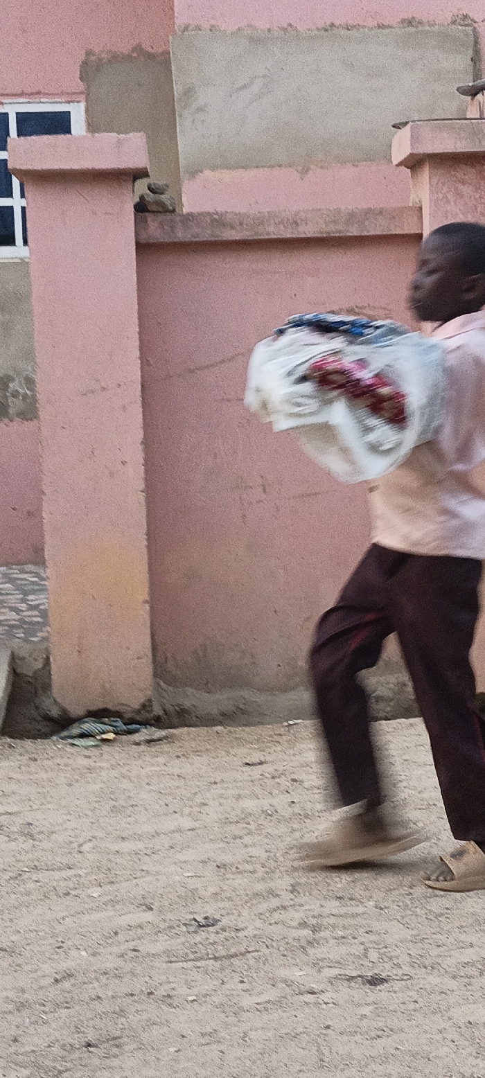 The image depicts a motion-blurred young person, likely a boy, walking or running outdoors in what appears to be a residential or urban setting. The boy is seen in profile, moving from right to left, wearing a light-colored, possibly pink or light orange, long-sleeved shirt and dark maroon or brown trousers with a lighter stripe down the side. He has light-colored sandals on his feet.
He is carrying a substantial bundle wrapped in white material, which appears to be plastic or cloth, and contains various colorful items, with hints of blue and red visible through the wrapping. The blur around the bundle and the boy's legs indicates movement.
The background consists primarily of a building or wall painted in a faded pink or peach color. Sections of exposed, unrendered light grey concrete are visible on the wall, showing areas where plaster might have been applied or is missing. A partial view of a window with white frames can be seen on the far left. A prominent pink vertical pillar stands in the mid-ground. On a horizontal ledge behind this pillar, small, stacked stones are visible. The ground is a light brown, sandy, or dirt surface, and some discarded, colorful material, possibly fabric, lies near the base of the wall on the left.
The lighting suggests it is daytime, possibly early morning or late afternoon, with soft, diffused light. There is no visible text in the image. The location context is Rangaza, Nigeria.