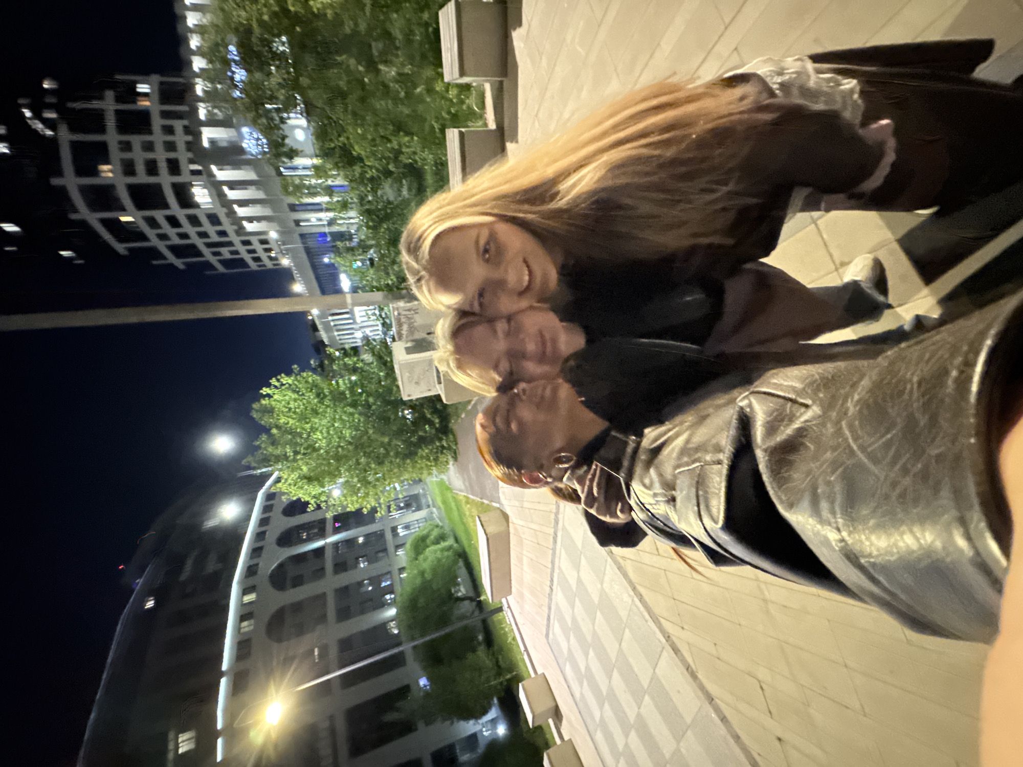 At night in Izhevsk, Russia, a selfie captures three individuals on a sidewalk near buildings and trees. Two women are closest to the camera: one with long blonde hair smiles, while another with short blonde hair rests her head on the first woman. The third person, with auburn hair, wears a black top and earrings. The ground is paved with light-colored tiles. In the background, buildings with illuminated windows stand tall, along with trees and streetlights.