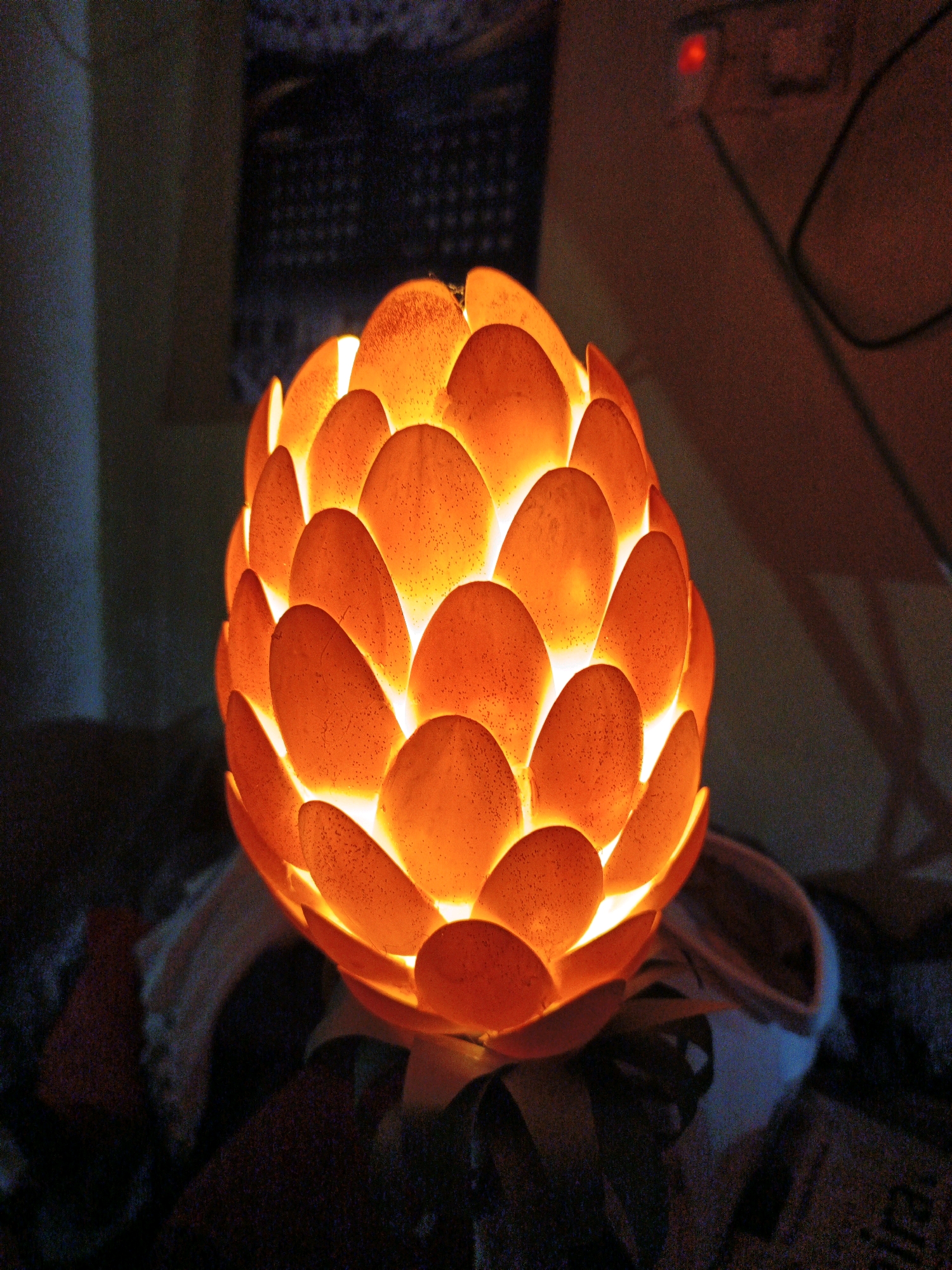 The image shows a close-up of an illuminated decorative lamp. The lamp is orb-shaped and covered in overlapping, petal-like segments that glow with a warm orange light from within. The texture of the segments appears to be slightly speckled. The lamp is positioned in a dimly lit environment, with hints of fabric or other soft materials visible in the foreground and background. To the right, a blurred background shows what appears to be a wall outlet with a red indicator light and a power cord. The overall impression is of a cozy, ambient setting. There is no indication of the city of Ketegan, Indonesia, in the image.