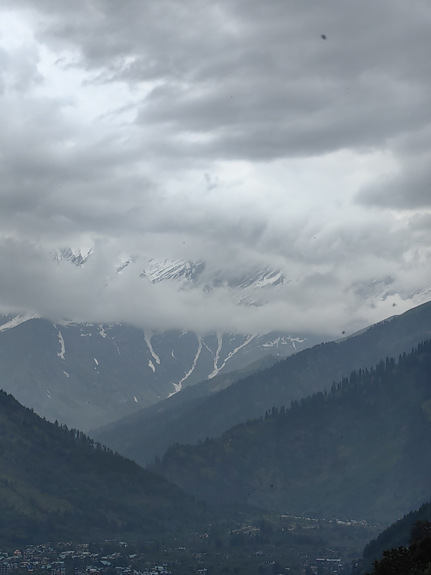 This is a photograph of a mountain landscape taken in the city of Vashisht, India. The sky is overcast with heavy, gray clouds. In the middle ground, snow-capped mountains are visible, with some peaks obscured by clouds. The lower slopes of the mountains are covered in dark green coniferous trees. In the foreground, a small town with buildings featuring green roofs is nestled in the valley. The overall impression is a cloudy day in a mountainous region.
