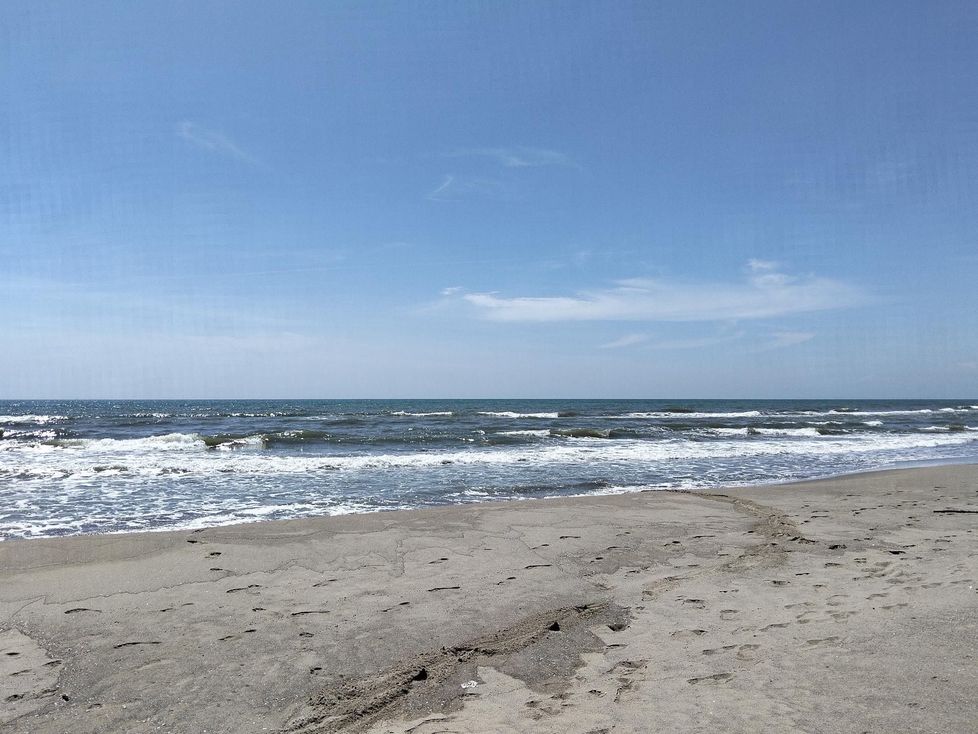 This media file captures a serene daytime view of a sandy beach in Mondragone, Italy, bordering the sea under a clear blue sky.
The upper half of the image is dominated by a bright, light blue sky with only a few faint, wispy white clouds scattered across it, suggesting fair weather. The sun is high, illuminating the scene brightly.
Below the sky, the horizon line separates it from the vast expanse of the sea. The water appears a deep blue-green, with numerous white-capped waves breaking and rolling towards the shore. The surface of the water glistens and reflects the sunlight, creating sparkling patterns. Closer to the beach, the waves are more pronounced, forming a band of white, foamy surf along the waterline.
The foreground is composed of light greyish-brown sand, which is wet near the water's edge and drier further up the beach. The sand is imprinted with numerous footprints, indicating that people have walked along this stretch of beach recently, although no individuals are currently visible. There are also subtle textures and lines in the sand, possibly from the retreating tide or previous activity. Small specks and shells are scattered across the sand, particularly closer to the water.
No people, structures, or visible text are present in the image. The scene conveys a sense of natural beauty and tranquility on a sunny day at the Italian coast.