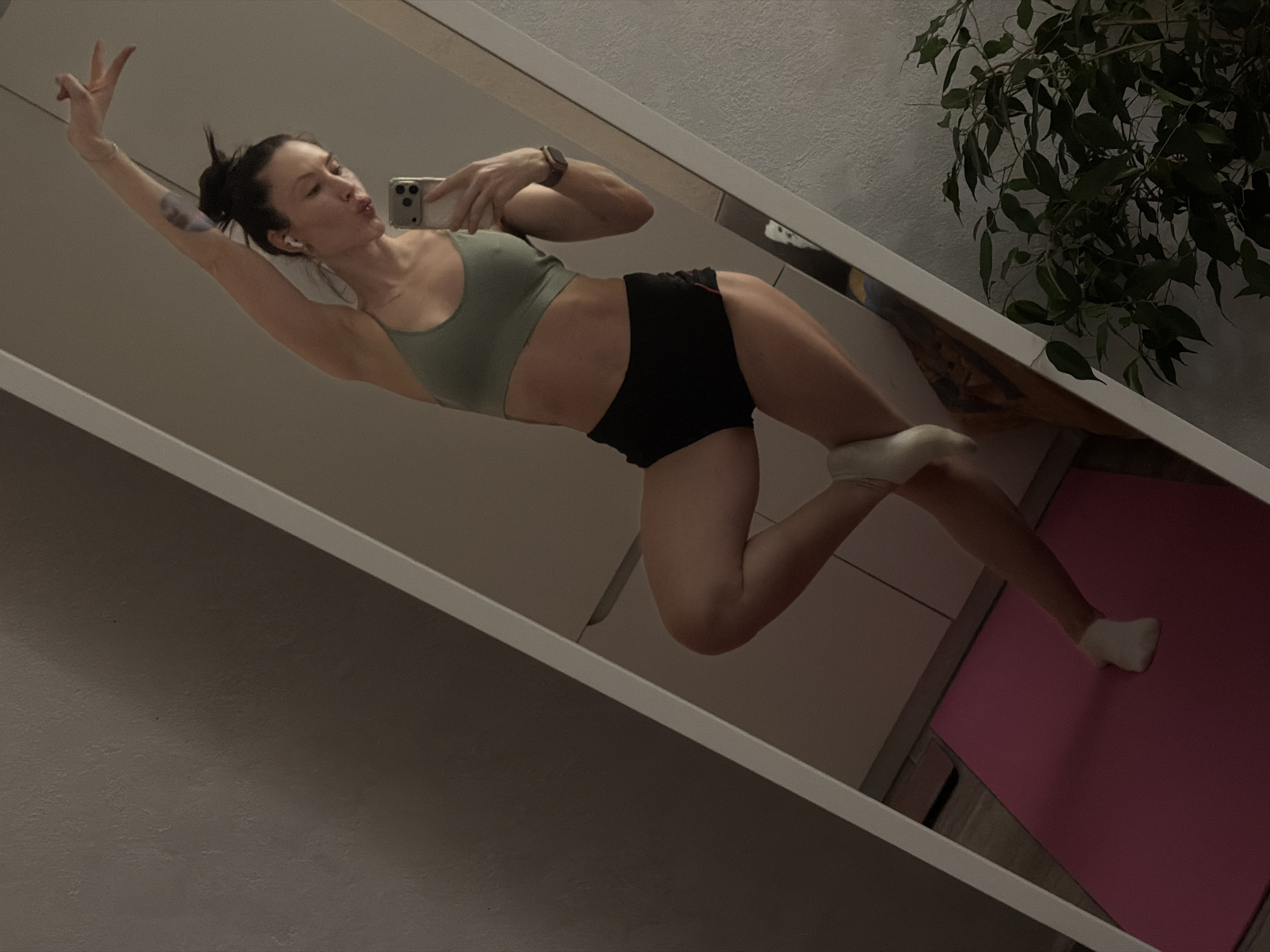 A woman is reflected in a mirror, appearing to be in an indoor setting. She is wearing a light green sports bra and black shorts, posing with a peace sign and pursed lips while holding a smartphone. Her hair is styled in a bun. To the right of the mirror, a green leafy plant is visible. A pink yoga mat is on the floor, partially visible in the mirror's reflection. The location is likely an apartment in Perm, Russia.
