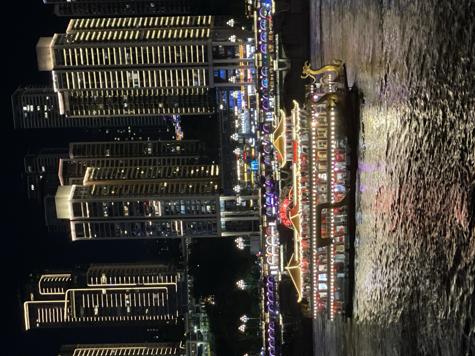 The image captures a vibrant night scene in the Yuzhong District of China, likely along the Yangtze River. In the foreground, a brightly lit, ornate boat with traditional Chinese architectural elements glides across dark, rippling water, its numerous lights reflecting on the surface. The boat features golden dragon sculptures at its bow.
In the midground, a waterfront promenade is illuminated with a series of arched structures adorned with colorful lights, suggesting a lively entertainment or commercial area. Streetlights with distinctive globe-shaped fixtures are visible along the promenade.
The background is dominated by a dense cityscape of modern skyscrapers, their facades outlined with bright, horizontal bands of light, creating a striking contrast against the dark night sky. The buildings exhibit a variety of architectural styles, with some featuring prominent vertical lighting elements. The overall impression is one of urban dynamism and a celebration of light and architecture at night.