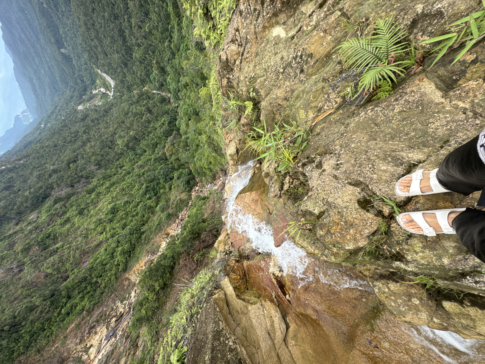 The image presents a high-angle view from a rocky cliff overlooking a dense, forested mountain valley. In the immediate foreground, a person's feet, wearing white sandals and dark trousers, are visible on the rugged, earthy-toned rock surface. The person appears to be standing at the very edge of a small, cascading waterfall, which tumbles down the rock face to the left.
The waterfall's white, foamy water contrasts with the wet, reddish-brown and grey rocks over which it flows. Lush green vegetation, including ferns and various grasses, grows on and around the rocky outcrops, adding vibrant pockets of color to the scene.
Beyond the immediate cliff edge, the landscape plunges into a vast expanse of verdant forest, covering steep hills and mountains that recede into the hazy distance under an overcast sky. A distinct, winding road can be seen cutting through the dense forest in the mid-ground, providing a sense of scale and human interaction with the natural environment.
The scene depicts a natural setting during daytime, likely a cloudy or diffused light day given the soft lighting. The overall impression is one of observation and immersion in a dramatic mountainous landscape, characteristic of Nam Khanh Vinh, Vietnam. No discernible text is visible in the image.