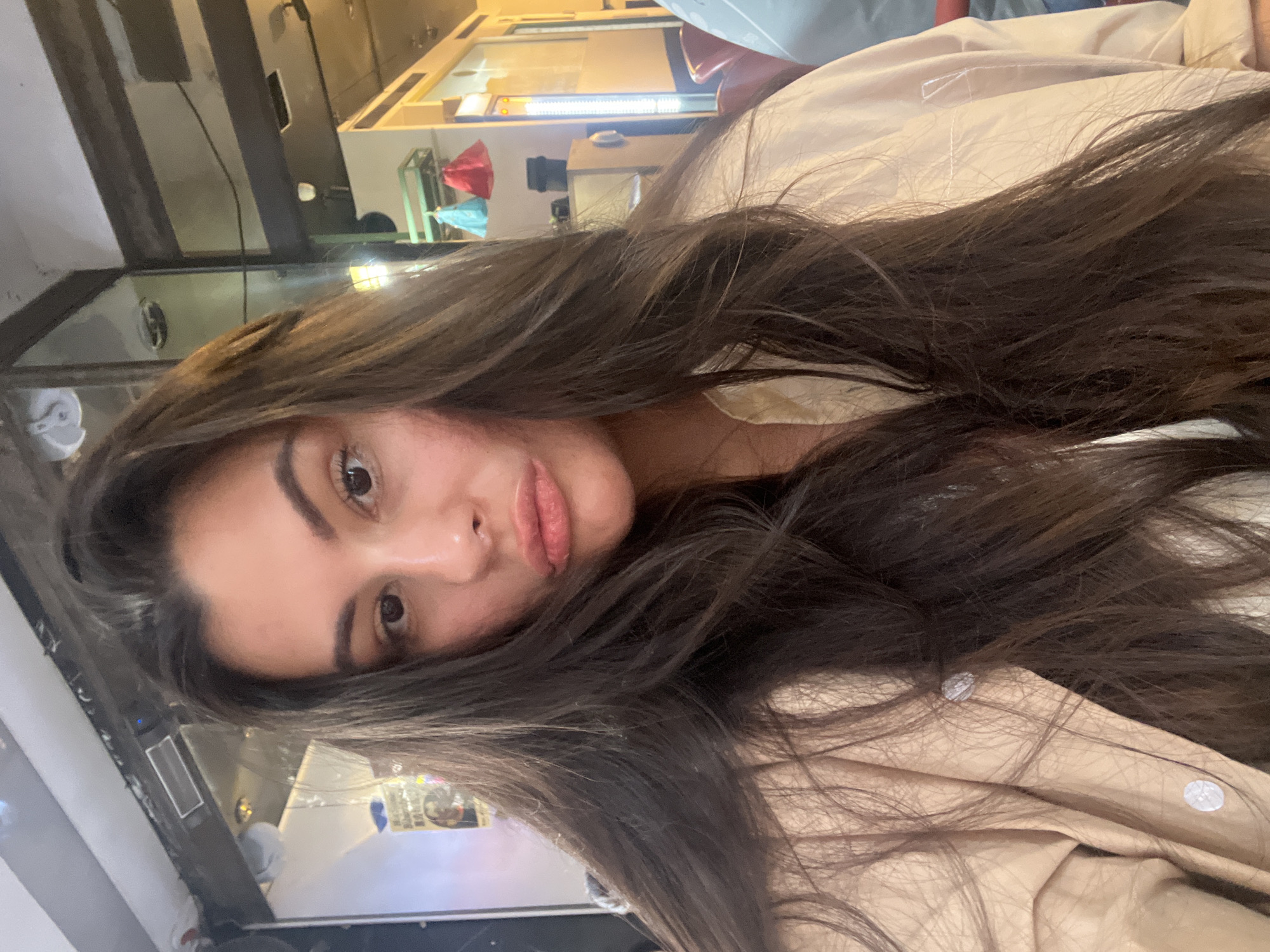 A close-up, selfie-style photograph features a young woman with long, dark hair, fair skin, and dark eyes. She is wearing a light beige or tan collared shirt or jacket, slightly unbuttoned at the top. Her hair is parted and falls loosely around her face and shoulders. Her expression is a subtle pout or duck-face. The background shows a blurred urban setting at nighttime. Through a window or opening, a glimpse of a brightly lit interior or storefront can be seen. There is no way to know that this photograph was taken in Tel-Aviv, Israel, based on the image alone.