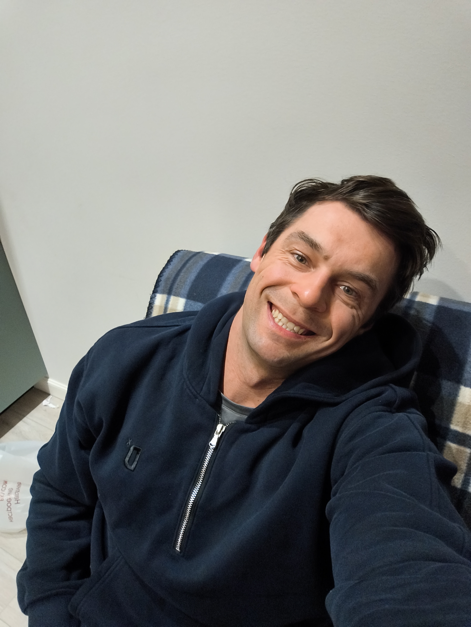 A medium, high-angle shot depicts a Caucasian male in a seated position, wearing a navy blue, zippered hoodie with a small, black logo on the upper left chest. He is smiling broadly with visible teeth. His hair is dark and slightly styled. Behind him is a blanket with a blue, gray, and tan plaid pattern. The background consists of a light gray wall. To the lower left, a portion of a light green object is visible along with a plastic bag with printed text.
There is no indication in this image that the location is Moscow, Russia.