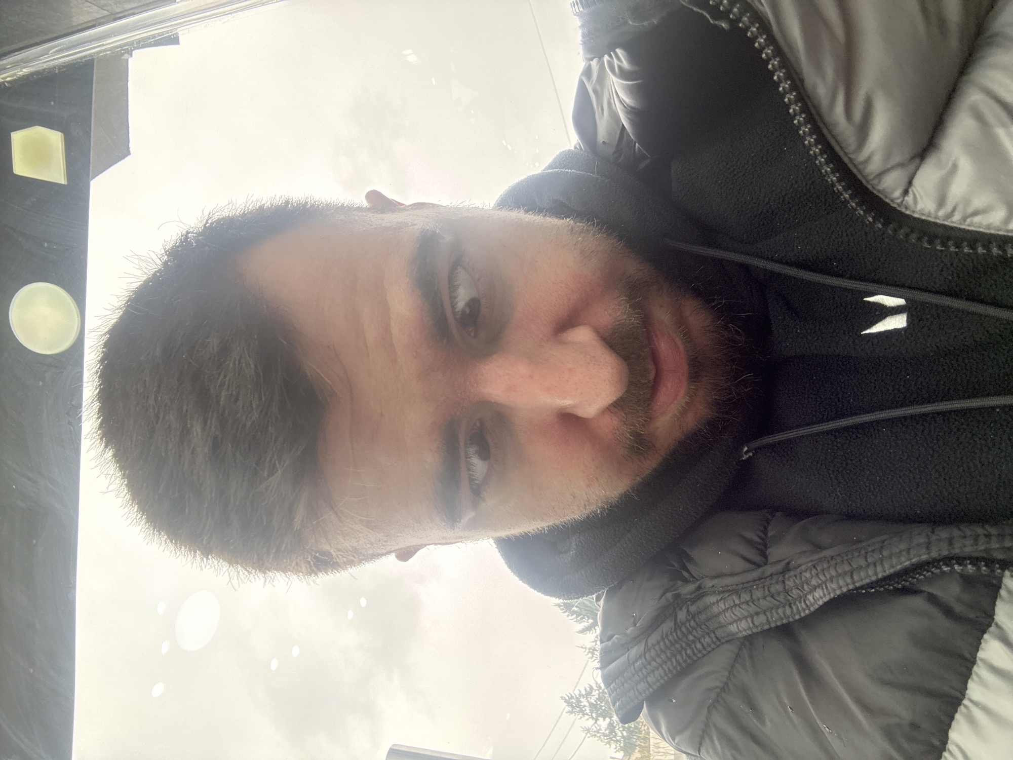 A man is looking into the camera. The man is wearing a jacket with a hood. The background is blurry, but it appears to be outdoors. The location is Sofia, Bulgaria.