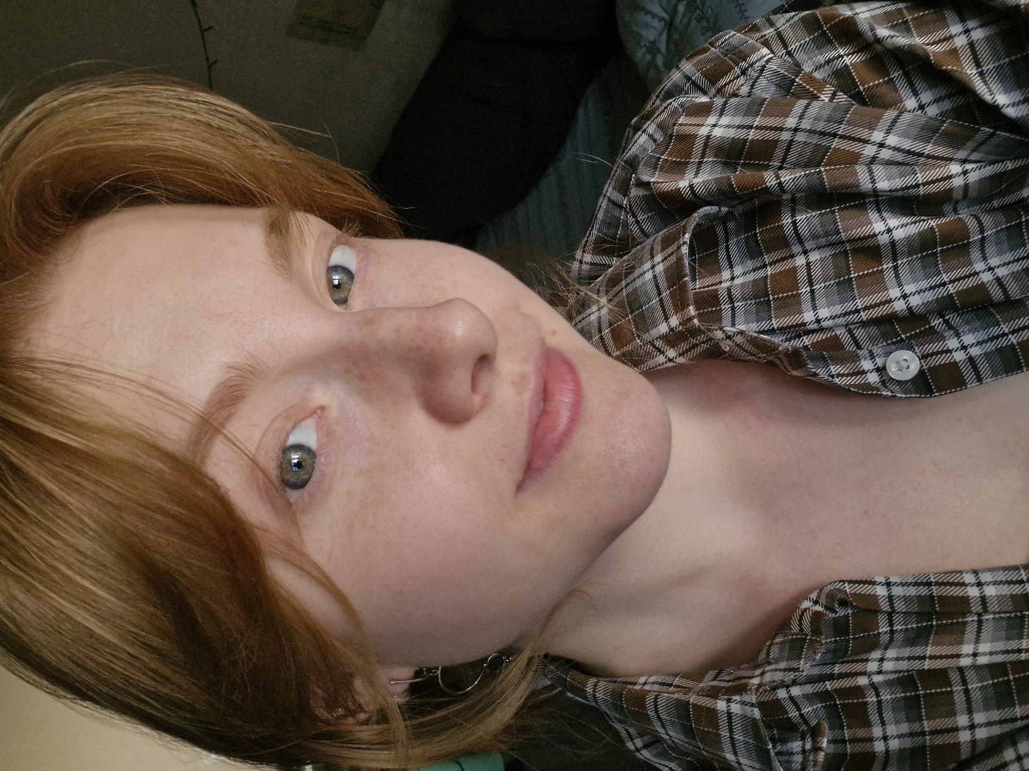 A selfie of a woman with red hair and freckles. She is wearing a brown and gray plaid shirt. Her eyes are light green. The background is blurry, showing what appears to be a dark-colored headboard. The location is not discernible from the given image.