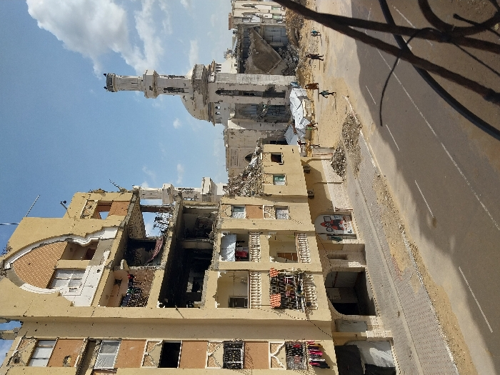 The image depicts a heavily damaged urban environment in Khan Yunis, Palestinian Territory, under a partly cloudy blue sky. In the foreground, a rusted metal railing is visible on the lower right, suggesting an elevated vantage point. A two-lane asphalt road with faded white markings runs across the mid-ground, bordered by a paved sidewalk on the left.
To the left of the road, a multi-story, light-colored residential building stands severely damaged. Multiple floors exhibit large sections of missing exterior walls, exposed interiors, broken windows, and visible rebar. An ornate arched structure is visible on one of the upper damaged sections. Laundry hangs from a balcony on one of the lower floors. The ground floor features arched entrances, and a partially obscured poster is visible on its facade. Piles of rubble and debris are scattered along the sidewalk and in the open areas beyond the road.
In the background, a large mosque complex with a central dome and a tall minaret shows significant structural damage, including cracks and missing sections. Several white, tent-like structures are visible among the debris near the mosque. Approximately 6-8 individuals, appearing to be civilians, are walking or standing amidst the rubble in the mid-distance between the road and the mosque, with some near the temporary shelters. No explicit interactions are discernable among the individuals; their action is primarily movement through the damaged landscape.
