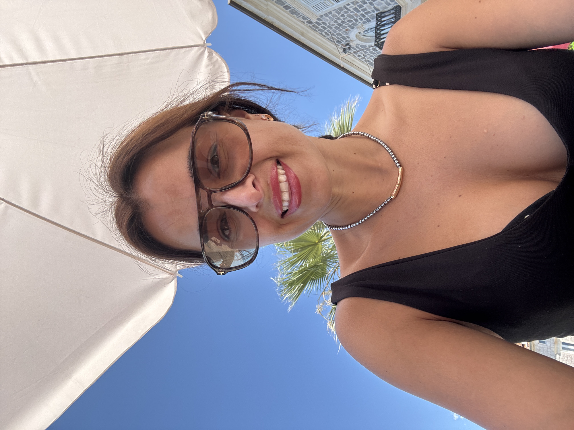 An outdoor selfie features a woman under a white umbrella. She is wearing large, brown-rimmed sunglasses and a black swimsuit. A beaded necklace with a gold bar pendant is around her neck. Her right shoulder and a portion of her chest are visible. In the background, a clear blue sky is visible, along with a palm frond and a building with stone construction and shutters. The overall scene suggests a warm, sunny day, possibly in a resort or coastal area.