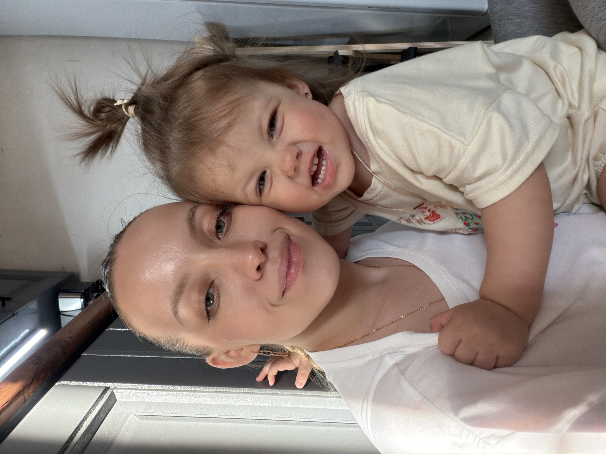 The image depicts a close-up indoor portrait of two females, an adult and a toddler, in Reshetnikovo, Russia. The adult female, positioned on the left, has fair skin, light-colored hair pulled back, and a subtle smile. She wears a white crew-neck t-shirt, a thin gold-colored chain necklace, and a gold-colored link-style drop earring in her left ear. Her left arm is around the toddler, with her hand faintly visible near the toddler's back.
The toddler female, positioned on the right and slightly lower, has fair skin and light brown hair styled with a high ponytail secured by a light-colored tie, and two smaller side ponytails. She exhibits a grimacing facial expression, with squinted eyes and an open mouth revealing upper teeth. She wears a light yellow or cream-colored short-sleeved t-shirt with a faint graphic design on the left chest and a thin gold-colored chain necklace. A small purple stud earring is visible in her right ear. Her right hand is clenched in a fist, grasping the adult's white t-shirt on the left chest area.
The background elements are out of focus. Behind the adult's left side is a light gray door or wall panel with vertical molding. Above the adult's head, a horizontal wooden beam is visible, with a modern linear LED light fixture mounted above it. Behind the toddler's right side, a light-colored wall is present, with faint dark, vertical elements and a gray upholstered surface, likely a sofa or chair, visible to the far right. The scene is brightly lit, with light originating from the left, casting some highlights and soft shadows.
