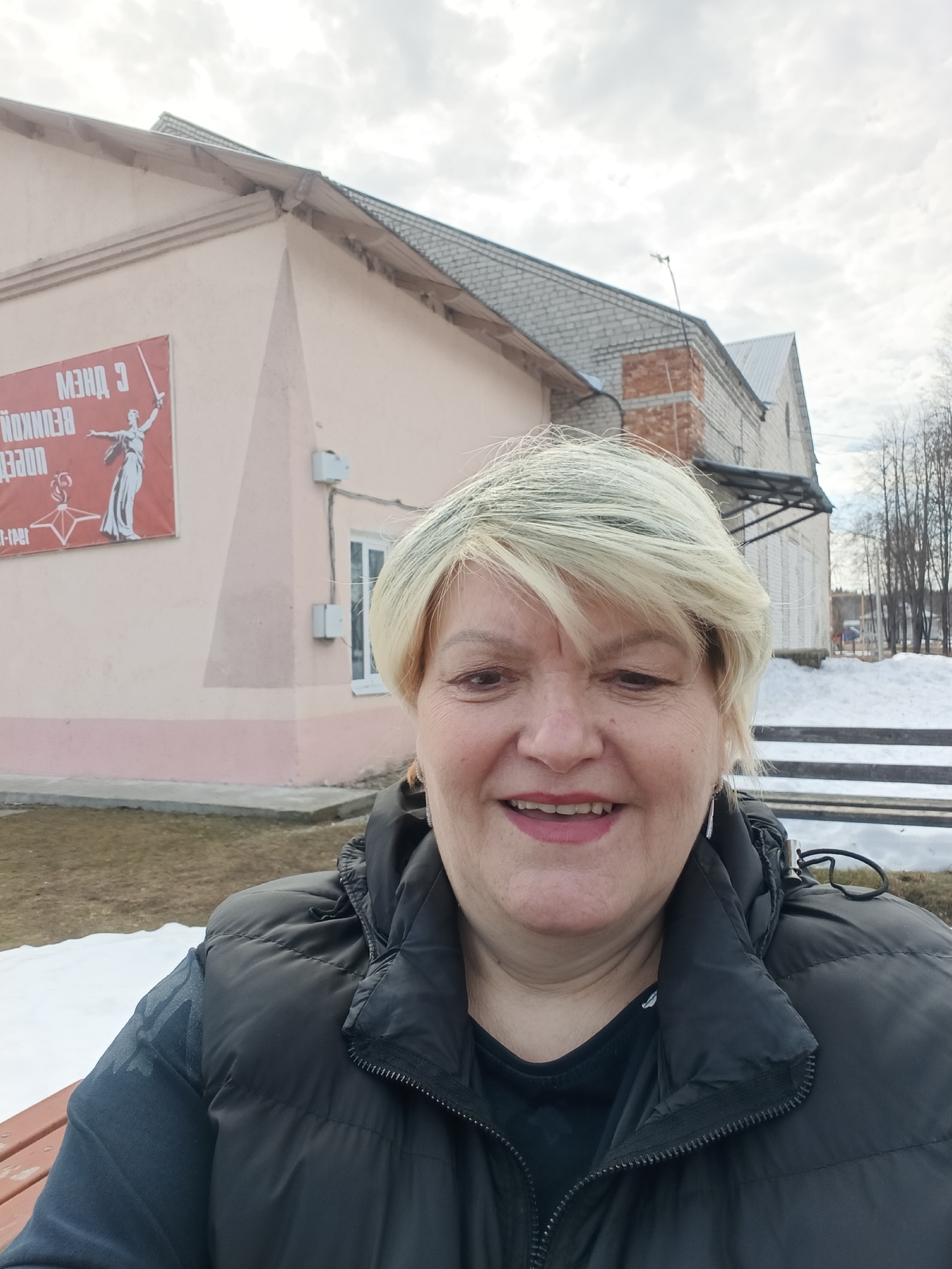 A middle-aged woman with short, blonde hair is taking a selfie outdoors in Красный Маяк, Russia. She is wearing a black puffer vest over a dark shirt. Behind her, there are two buildings. The first is a pale pink building with a red banner depicting a woman holding a sword. The banner reads "МЭИД С." A window and electrical boxes are visible on the side of the building. The second building is made of light-colored bricks with a metal roof. There is snow on the ground, indicating it is likely winter or early spring. Bare trees are visible in the background.
