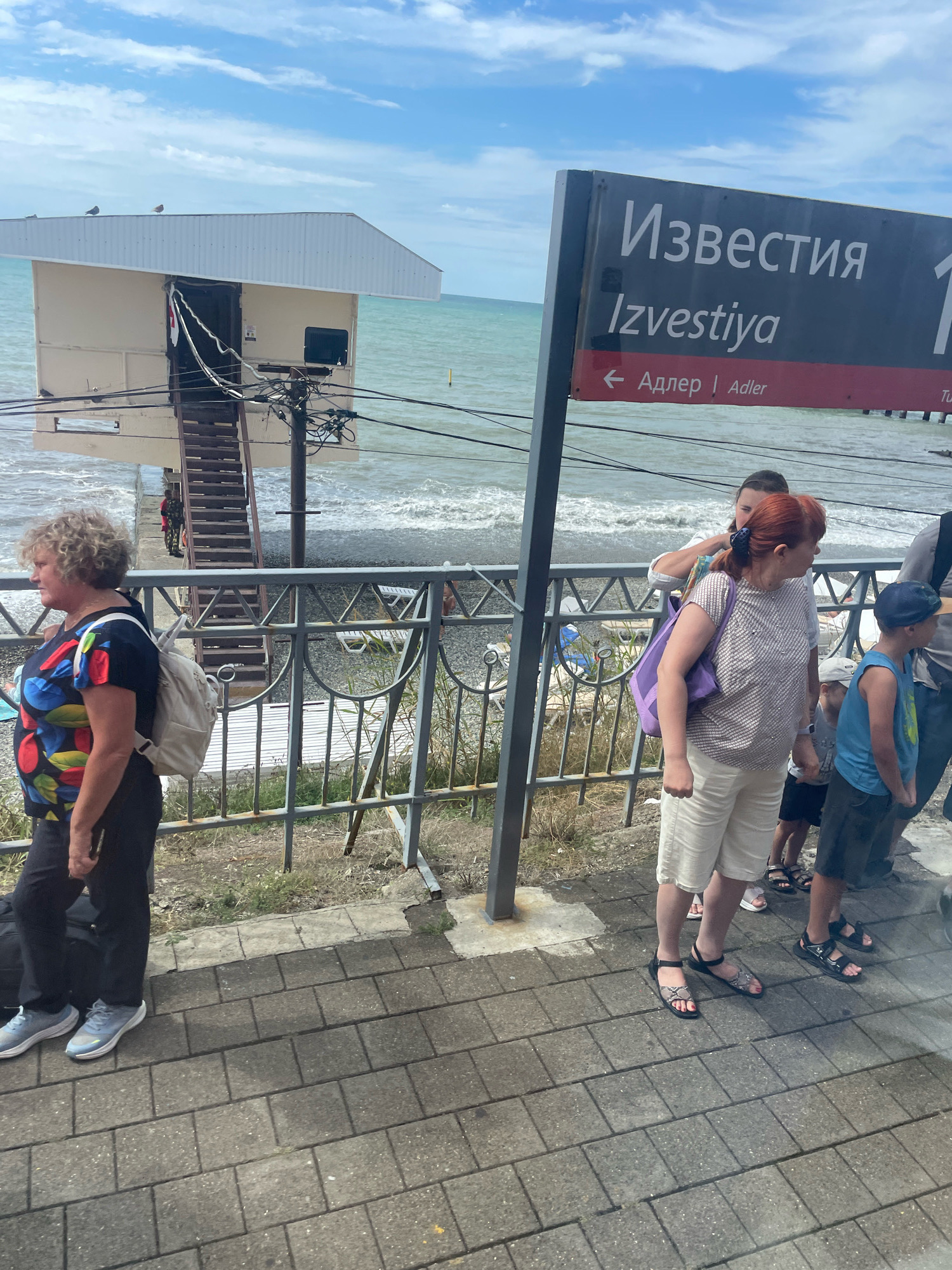 The image shows a signpost with Russian and English text, indicating "Izvestiya" and directions to "Adler". The sign is positioned near a body of water, likely the Black Sea, with a beach and a small building on stilts in the background. Several people are standing on a paved area in front of a railing, some with luggage. The overall scene suggests a coastal location, possibly a train station or a public area near the sea, within the city of Sochi, Russia. The weather appears to be overcast.