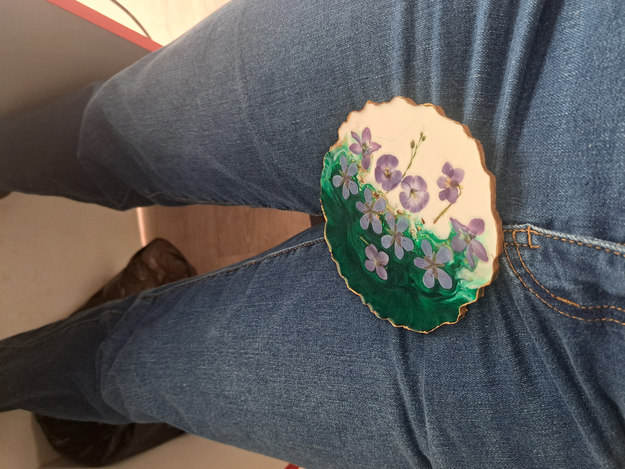 A top-down view shows a person wearing blue jeans with a round, decorative coaster resting on their right thigh. The coaster has a white background, green accents, and purple flower designs. A small portion of a brown shoe is visible in the lower left corner, along with a glimpse of a table. The scene appears to be indoors. There is no evidence to suggest this is in Leningradskaya, Russia.