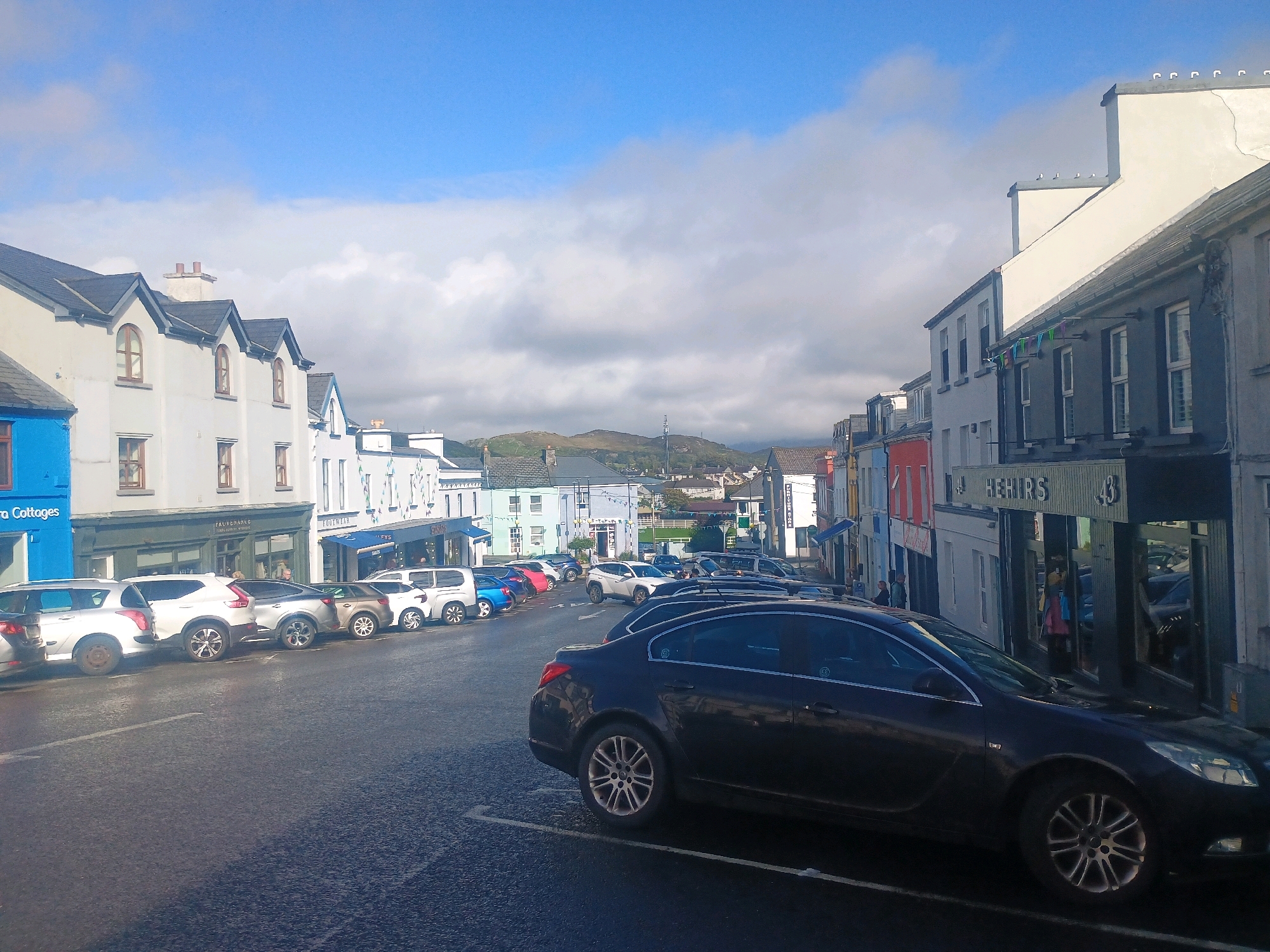The image depicts a street scene in what appears to be a coastal town. In the foreground, a dark-colored sedan is parked at an angle. The street is paved and lined with parked cars, predominantly SUVs and smaller vehicles. On either side of the street are buildings, many of which appear to be shops or residences, with colorful facades. Some buildings have awnings and signage, suggesting commercial activity. In the background, rolling hills under a partly cloudy sky frame the town. The overall impression is of a quiet, everyday scene in a European town.