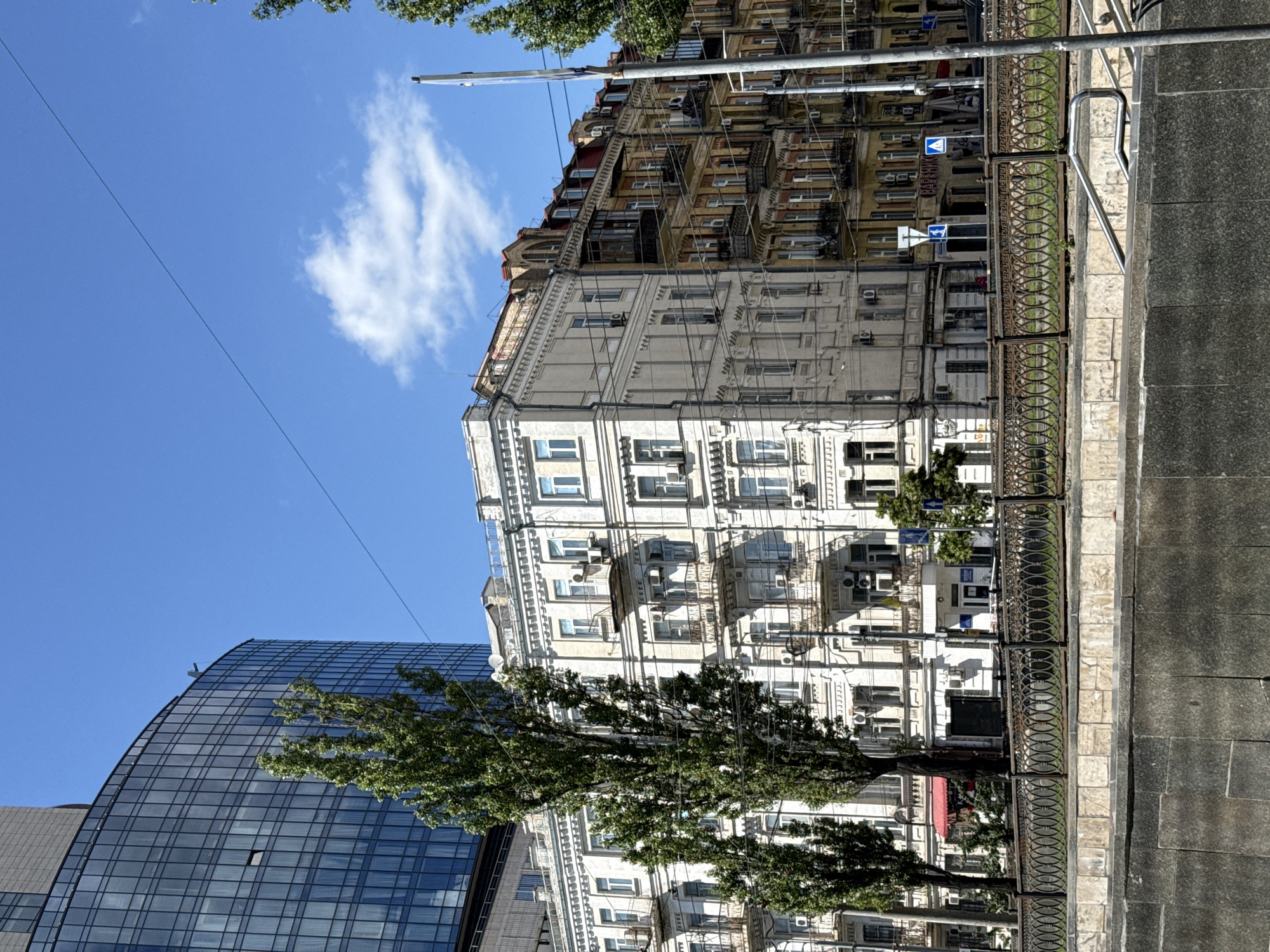 The image depicts a street scene in Kyiv, Ukraine, showcasing a juxtaposition of modern and historic architecture. On the left, a tall, modern glass-fronted building with a curved facade dominates the skyline. To its right, several older, ornate buildings with cream-colored and yellow facades line the street. These buildings feature balconies with intricate metalwork and arched windows. Tall, leafy trees are positioned in front of these structures, partially obscuring them. Overhead, power lines crisscross the sky. A metal fence with a decorative pattern separates the sidewalk from a lower level that appears to be a retaining wall. The sky is bright blue with a few scattered white clouds. The overall impression is of a vibrant urban environment with a rich architectural history.