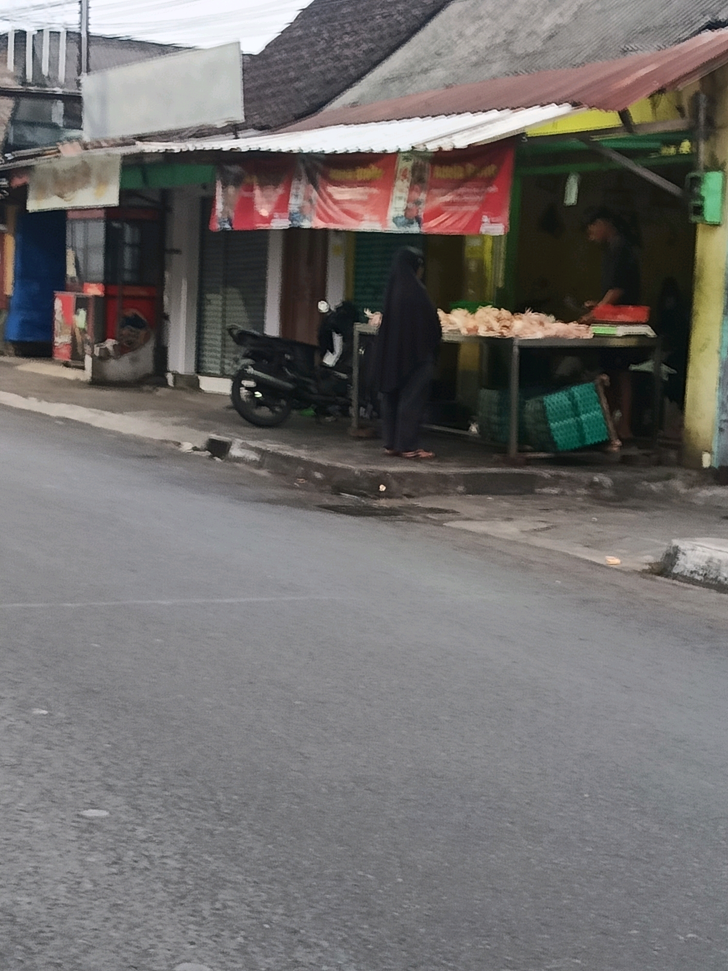 The image shows an outdoor market scene in Yogyakarta, Indonesia, with a focus on a food stall. A woman in a black hijab is standing on the left, facing a table laden with raw chickens. To her right, a man in a dark shirt is working at the stall, possibly preparing or selling the chicken. The stall itself is covered by a red and white banner advertising food. Several green plastic crates are visible beneath the table, and a black motorcycle is parked nearby. The market appears to be located along a street, with buildings and a rooftop visible in the background. The overall lighting suggests an overcast day or early morning.