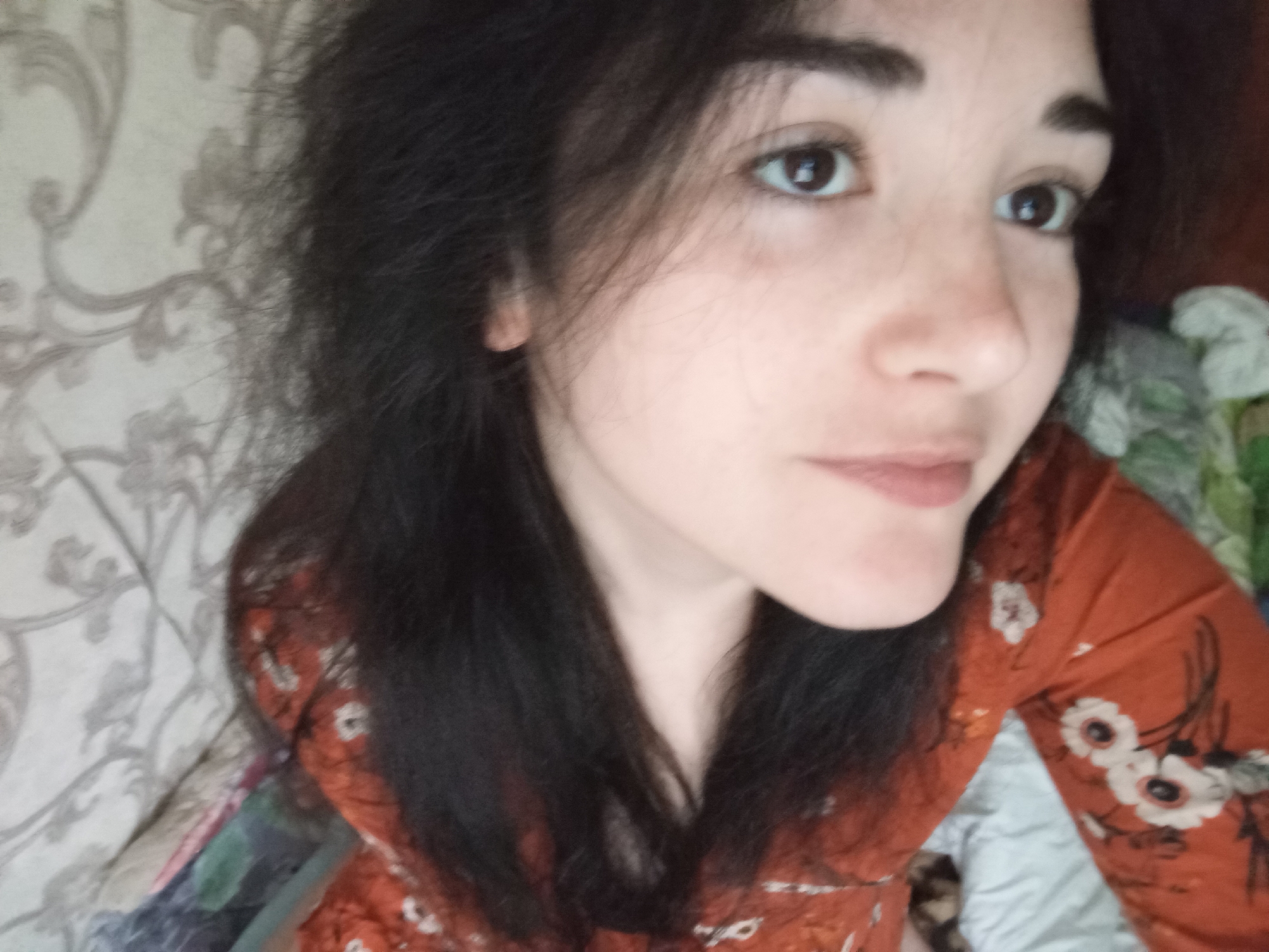 A close-up shot shows a young woman with dark hair, brown eyes, and fair skin. She is wearing an orange floral dress. The background includes a patterned wall or wallpaper and possibly bedding. The image does not provide sufficient information to determine a specific location in Poltava, Ukraine. There are no discernable actions or interactions.