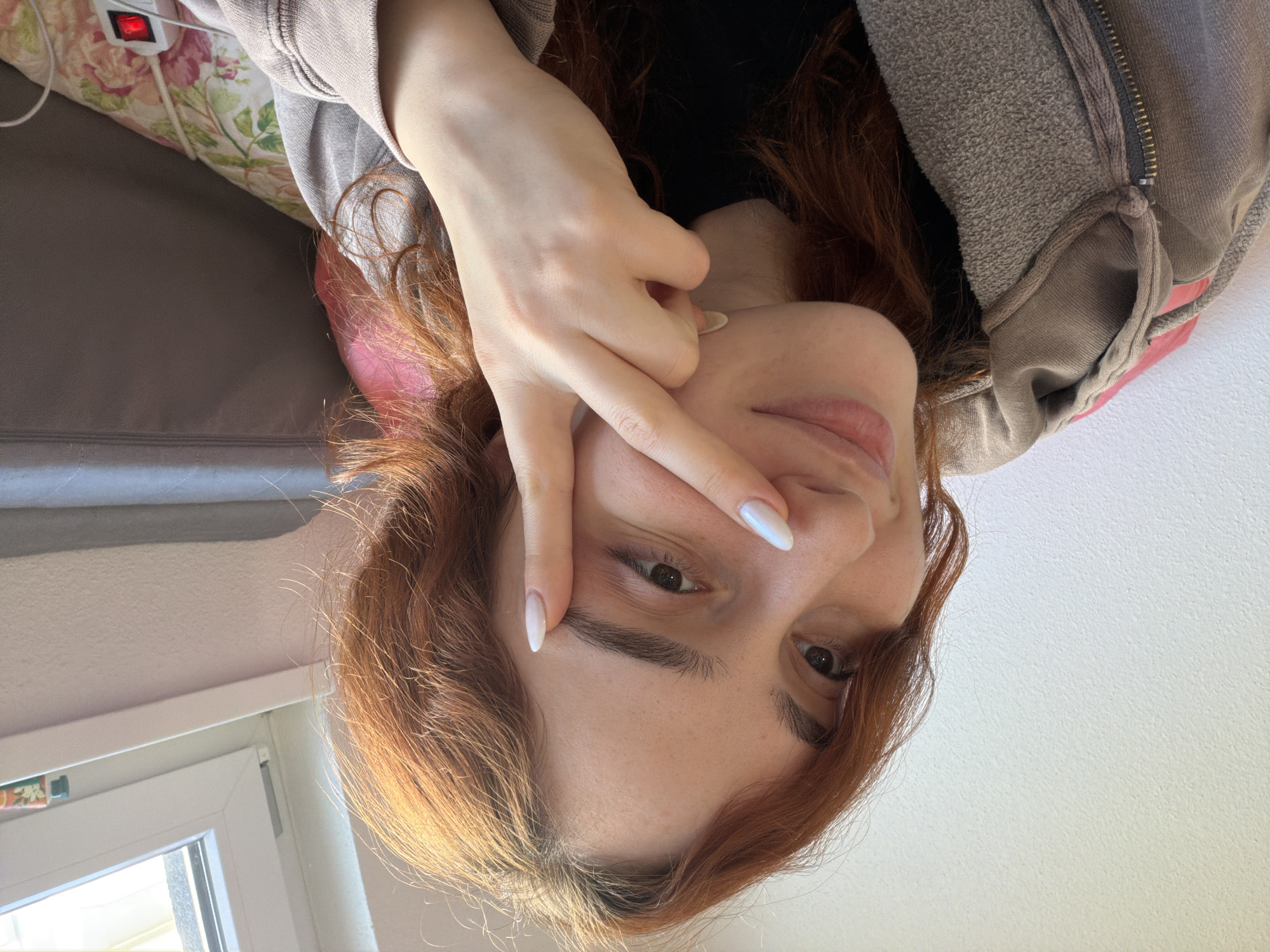 This inverted, close-up indoor shot, taken in Strasbourg, France, features a young woman with reddish-brown, wavy hair. She is looking directly at the viewer with a neutral, perhaps slightly playful, expression. Her left hand is raised, forming a "V" sign or peace gesture with her index and middle fingers, partially obscuring her left eye. Her fingernails are adorned with a light, possibly iridescent white polish.
The woman is wearing a light grey hooded sweatshirt or jacket, with details of its texture and a zipper visible on her right side. The background suggests a domestic setting, likely a bedroom or living room. Behind her, to the left, is a floral patterned pillow in shades of pink, green, and white. Resting on this pillow is a small white electronic device featuring a prominent red illuminated light, possibly a power indicator or button. Below the pillow, a section of a grey sofa or bed frame is visible. Further to the left and slightly behind her, a white window frame is present, allowing bright daylight to enter, indicating the photograph was taken during the day. The right side of the image shows a plain white wall. No visible text is present in the image.
