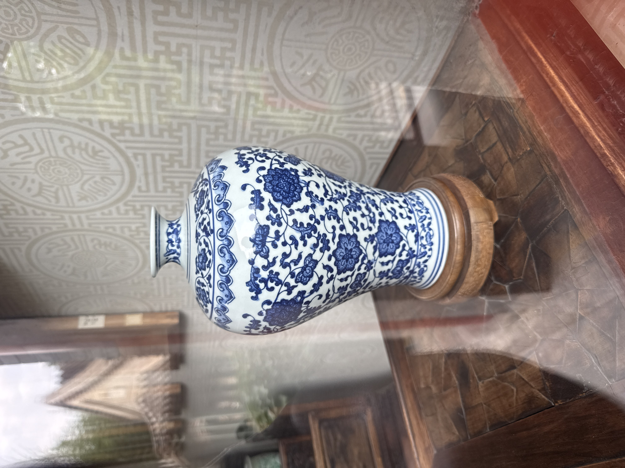 A close-up, eye-level shot captures a blue and white porcelain vase with intricate floral patterns, displayed on a polished wooden table in Xicheng District, China. The vase rests on a circular wooden stand, and its reflective surface mirrors the surrounding environment. The background features a textured wallpaper with a repeating circular motif and geometric patterns. To the left, a reflection in a glass surface reveals a glimpse of a traditional Chinese building with a gabled roof, surrounded by greenery. The scene is lit with ambient light, creating soft shadows and highlights on the objects and surfaces.