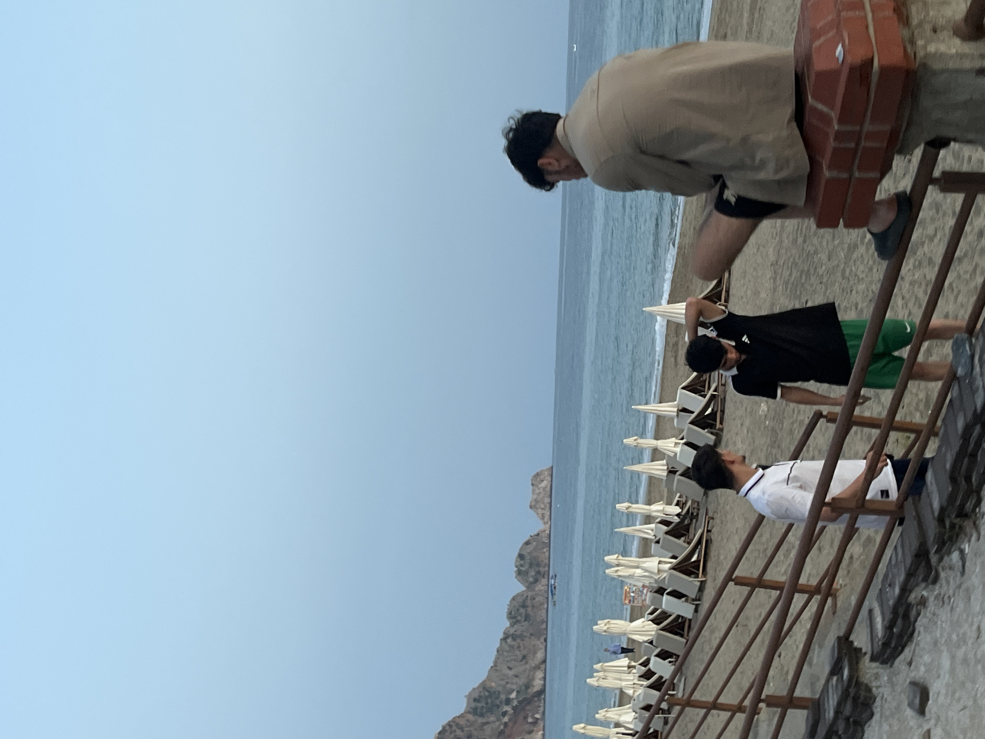 The image depicts a coastal scene at a beach in Alanya, Turkey, with several individuals present. A man in a beige shirt is leaning forward, possibly observing the scene. Below him, two other men are standing on a set of metal stairs leading down to the beach. The first is wearing a white shirt, while the second is in a black shirt and green shorts.
The background reveals a row of tightly packed beach umbrellas and sunbeds, indicating a tourist area. Beyond the umbrellas, the Mediterranean Sea stretches out, with a landmass or cliff visible on the horizon. The sky appears overcast, suggesting a somewhat cloudy day.